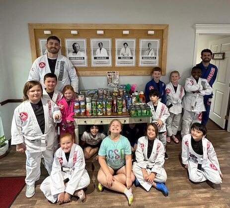 Group of children and adults in martial arts uniforms around a table with canned goods, indoors.