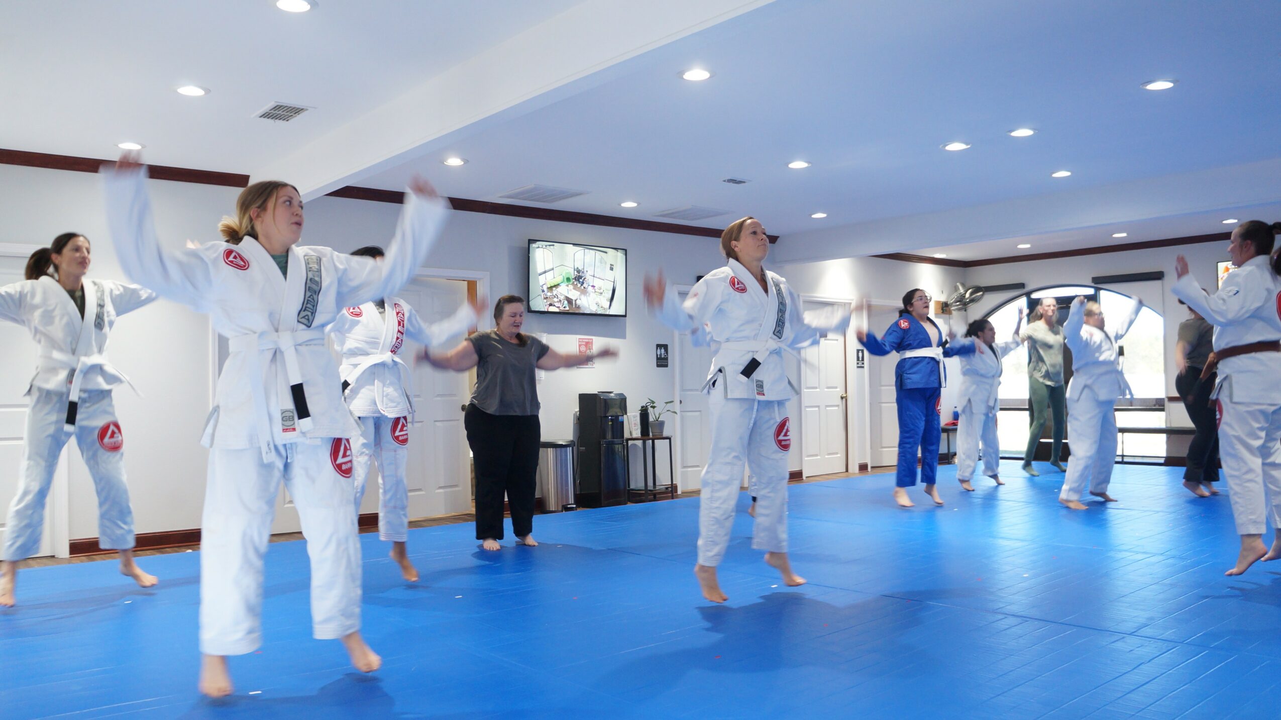 People practicing martial arts in a dojo with blue mats and white walls, some with arms raised, others observing.