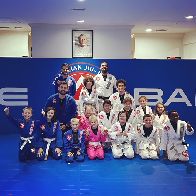 Group of children and adults in Brazilian Jiu-Jitsu uniforms posing on a blue mat, with a blue wall and a portrait above. 20 words.