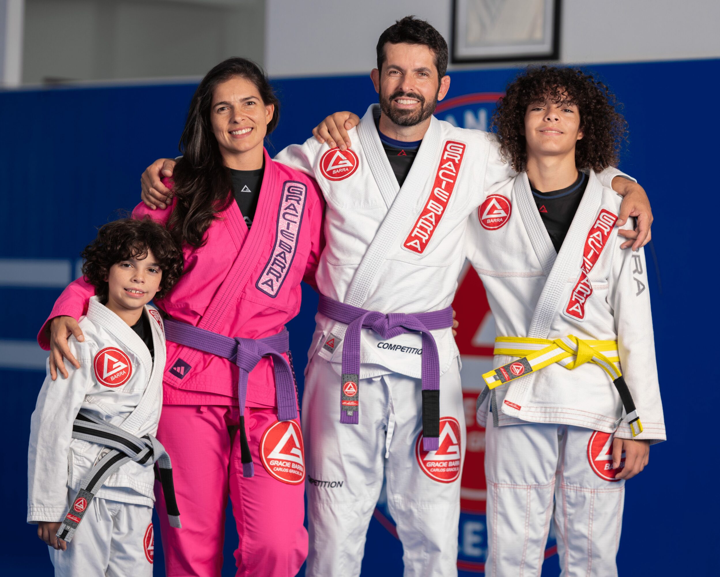 Four people in Brazilian Jiu-Jitsu uniforms smiling, standing together in a gym. Two children and two adults, one man and one woman.