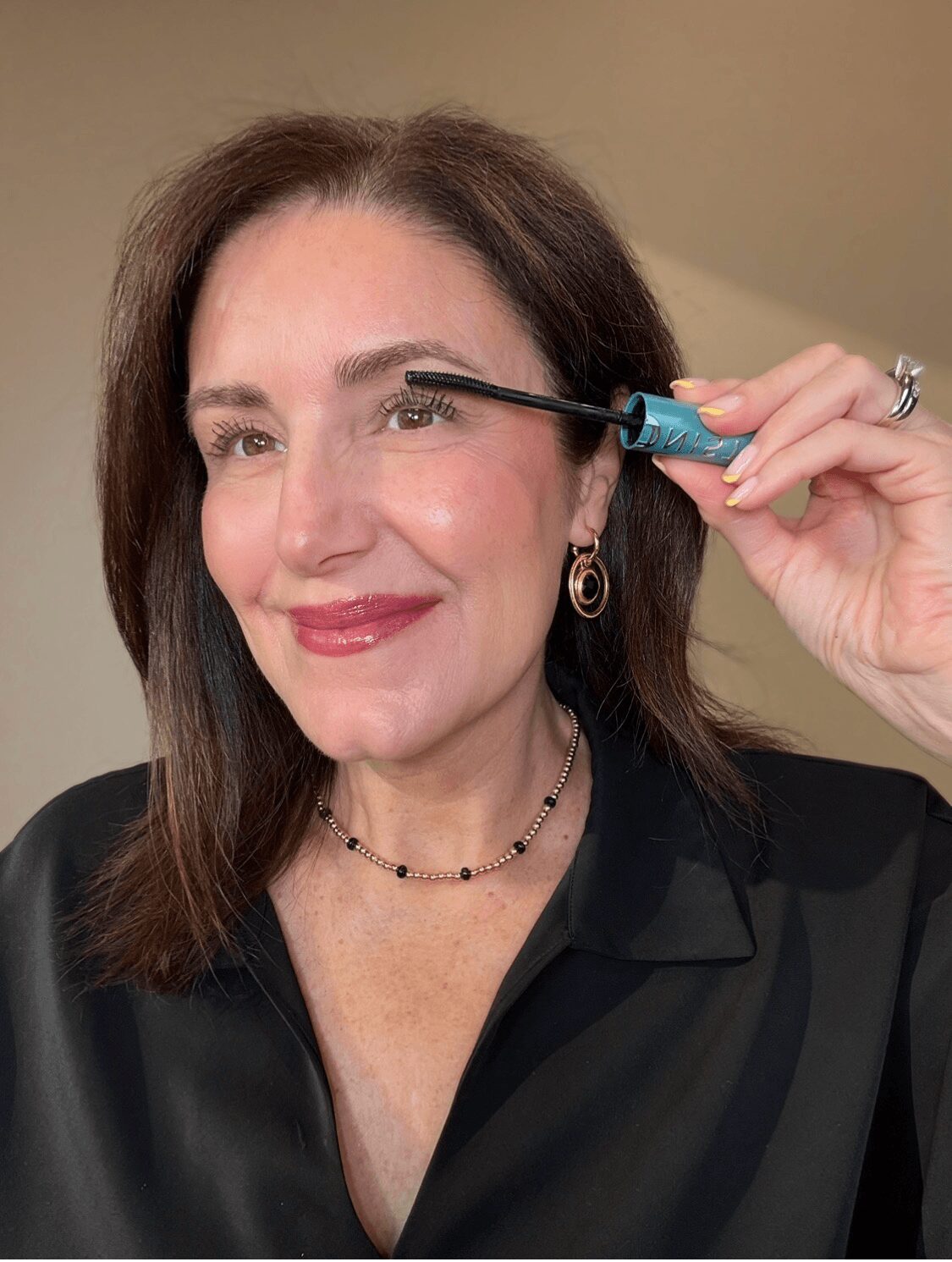 Woman applying mascara to her eyelashes, smiling, with shoulder-length dark hair, wearing earrings and a black top.