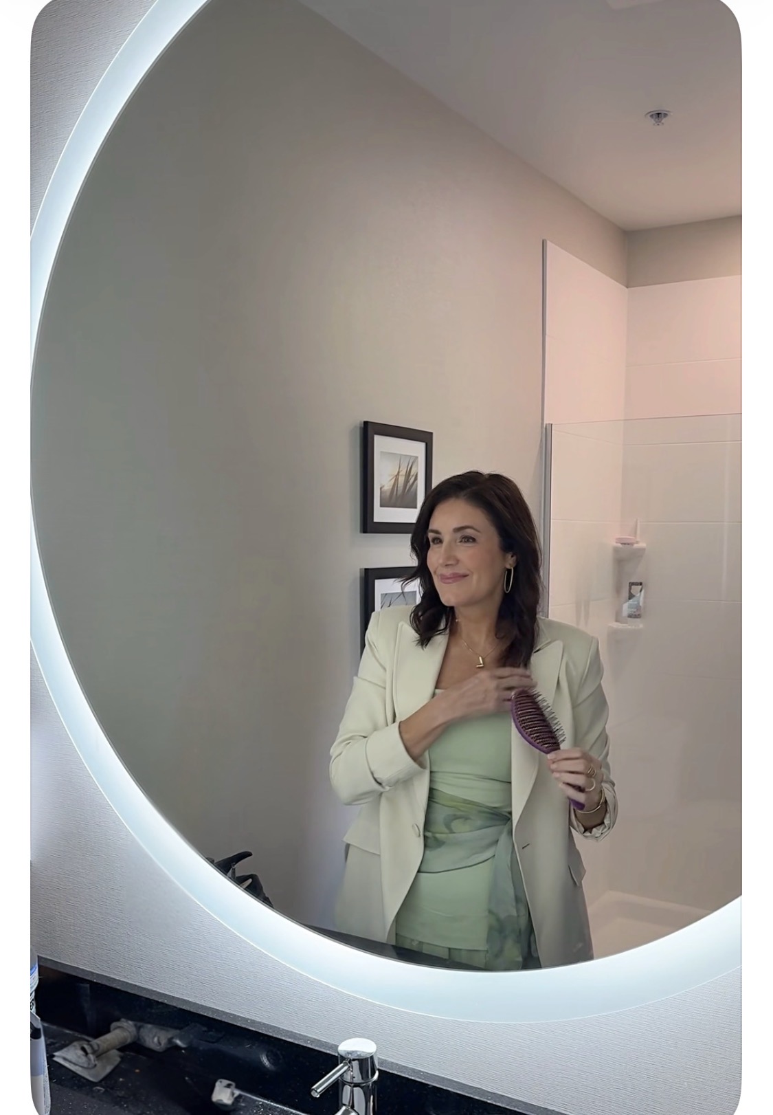 Woman taking a selfie in a mirror with a circular light frame, holding a phone, smiling, in a room with framed pictures.