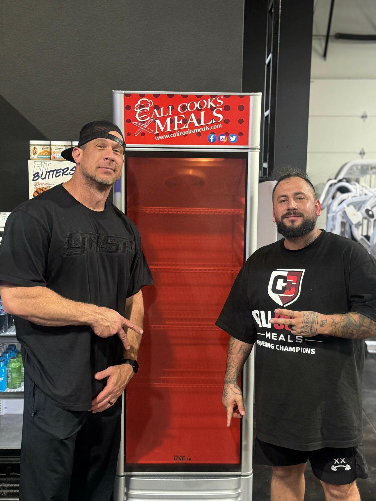 Two men standing in front of a red display shelf with a sign reading 'Cali Cooks Meals'.