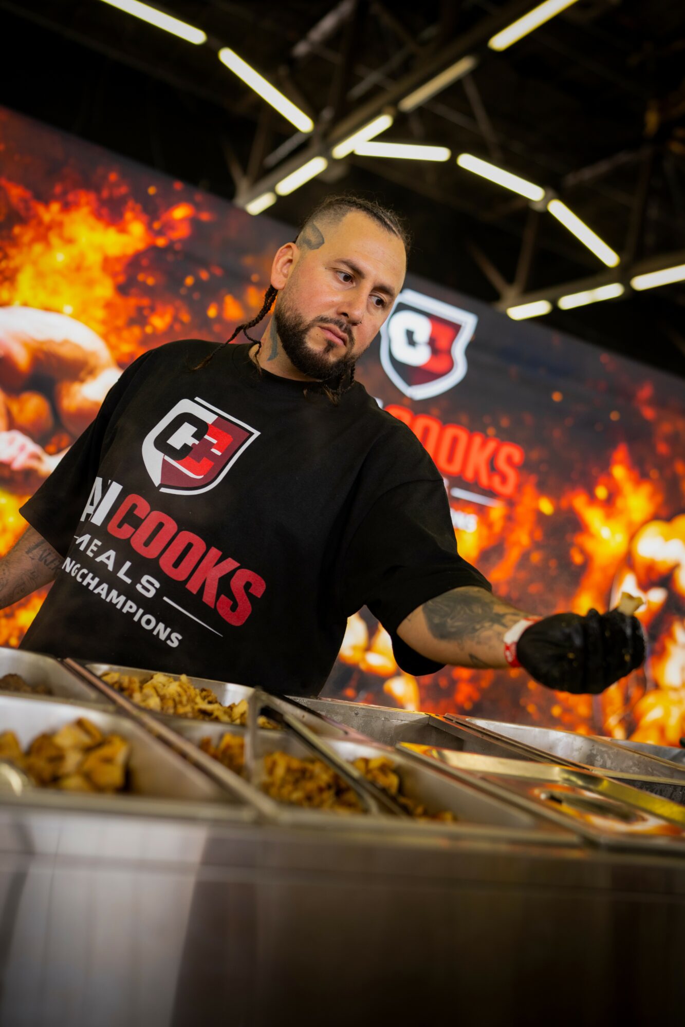 Man serving food at a buffet with a fiery background and logo, wearing a black shirt with red and white text.