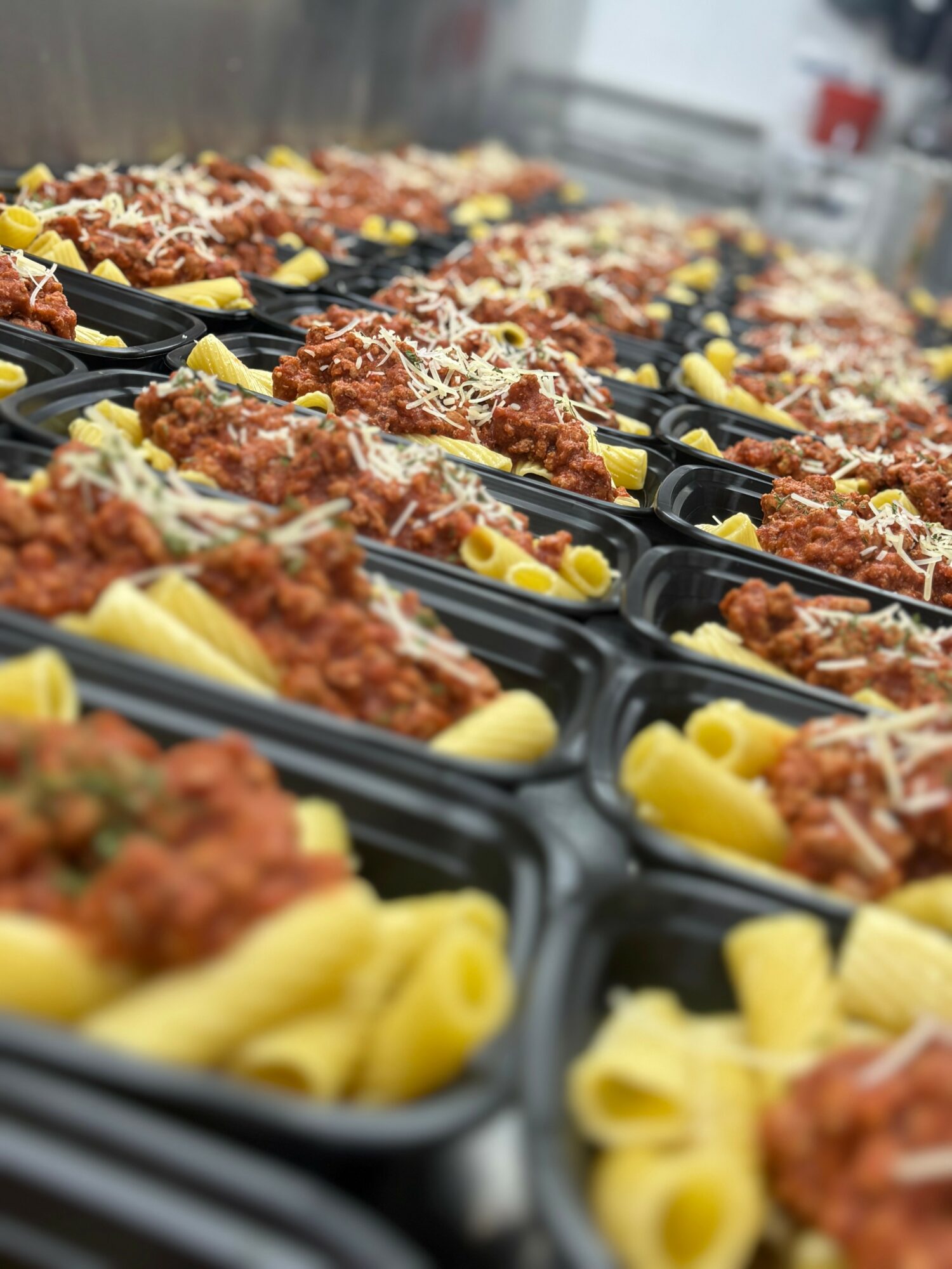 Multiple black meal trays filled with pasta, tomato sauce, grated cheese, and garnishes, arranged in rows on a table.