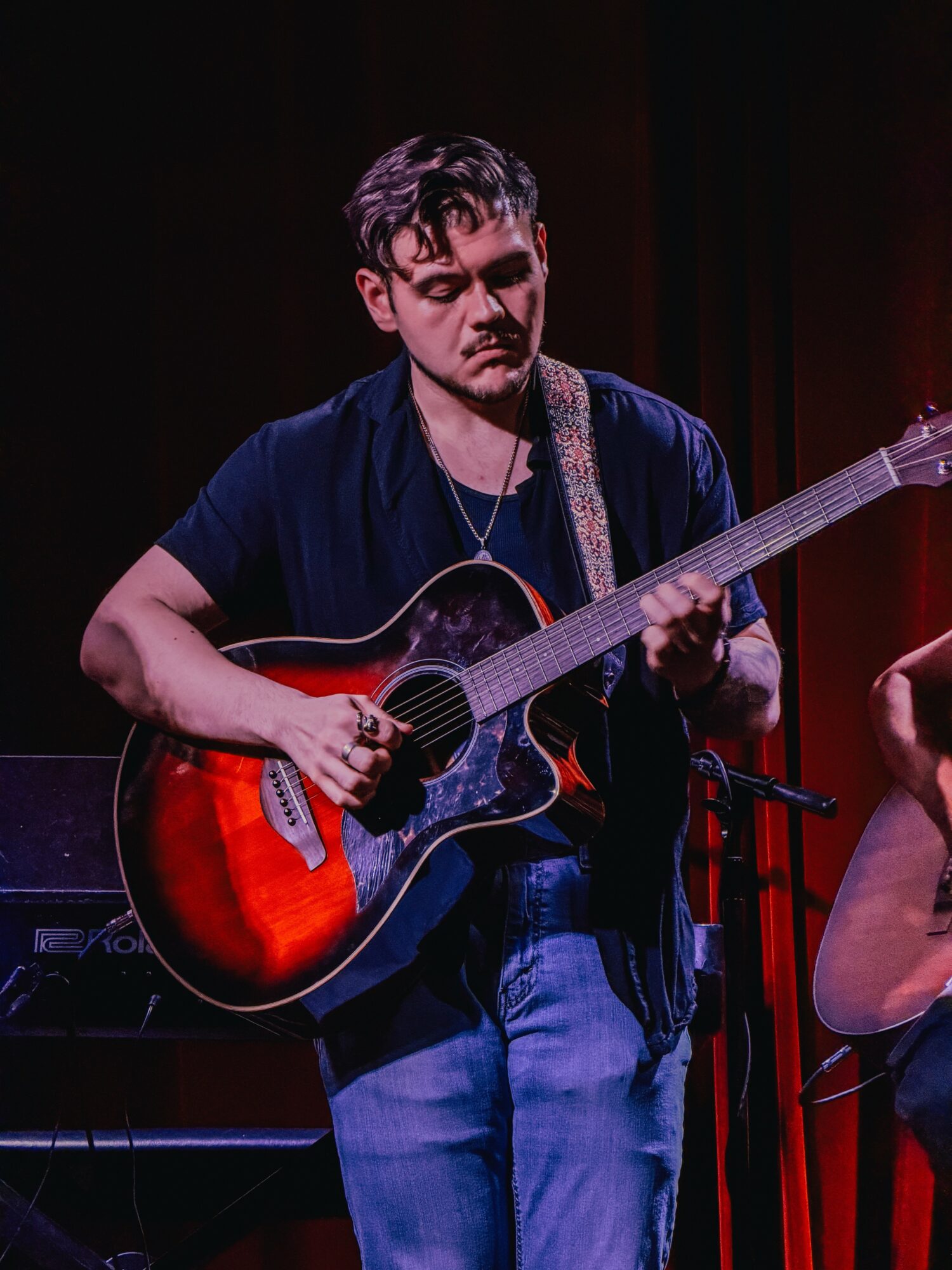 Young man playing an acoustic guitar on stage, wearing a dark shirt and jeans, with a red curtain background.