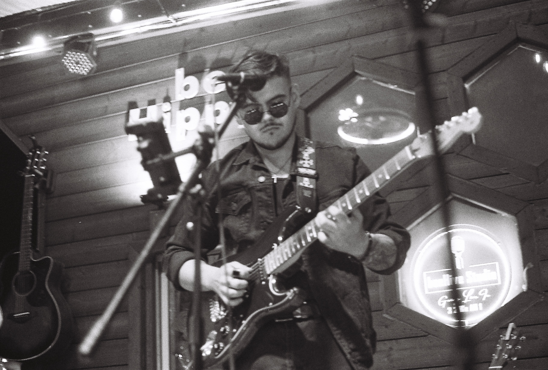 Person playing electric guitar on stage with wooden wall background, wearing sunglasses and a jacket.