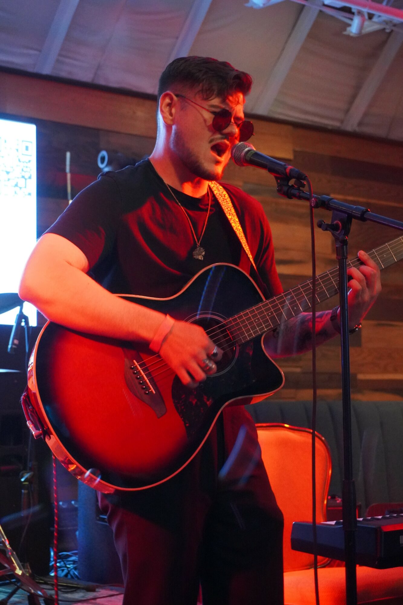 Person playing guitar and singing into microphone on stage with wooden background, illuminated by stage lights.