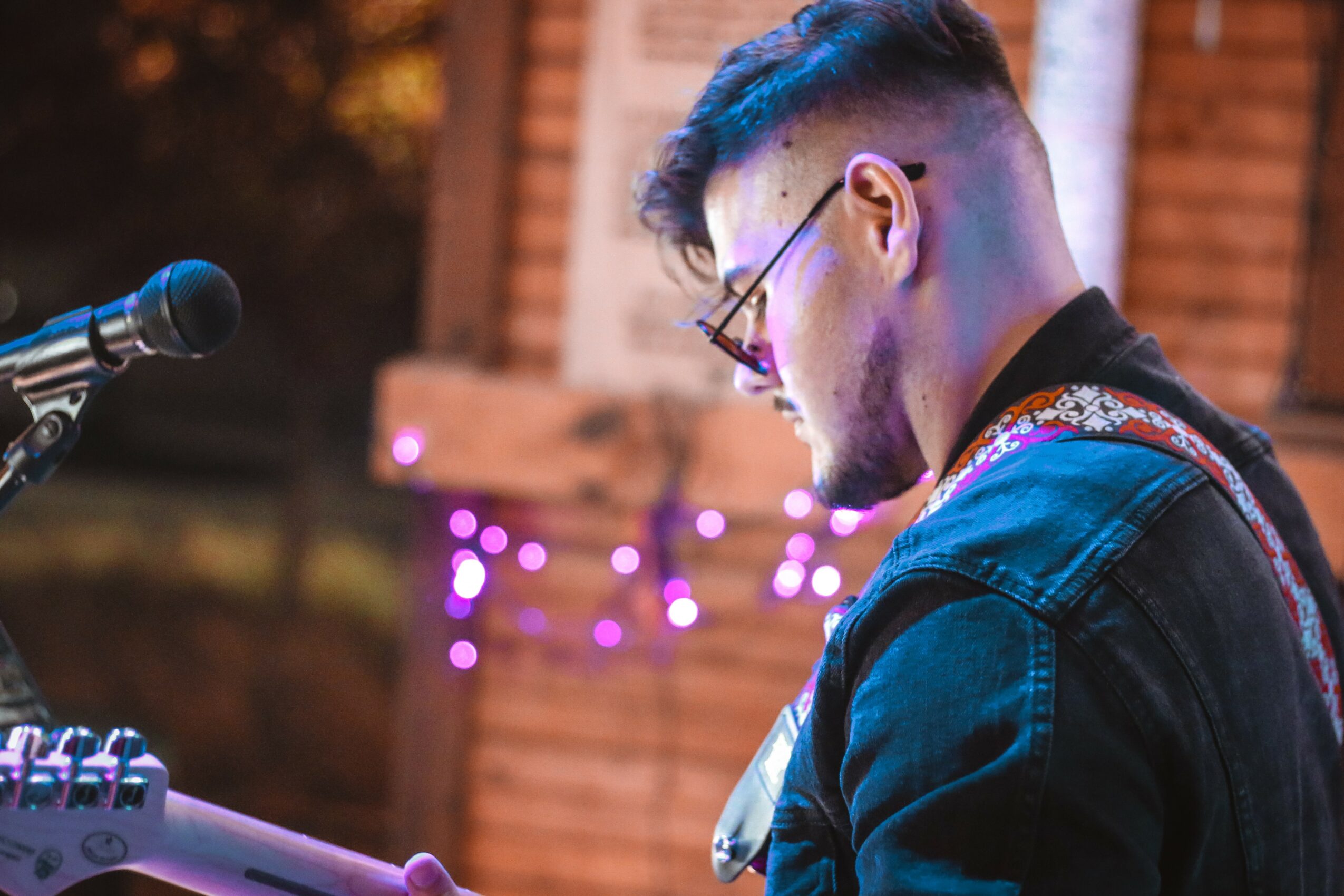 Young man with glasses and a beard playing music outdoors at night, with purple string lights in the background.