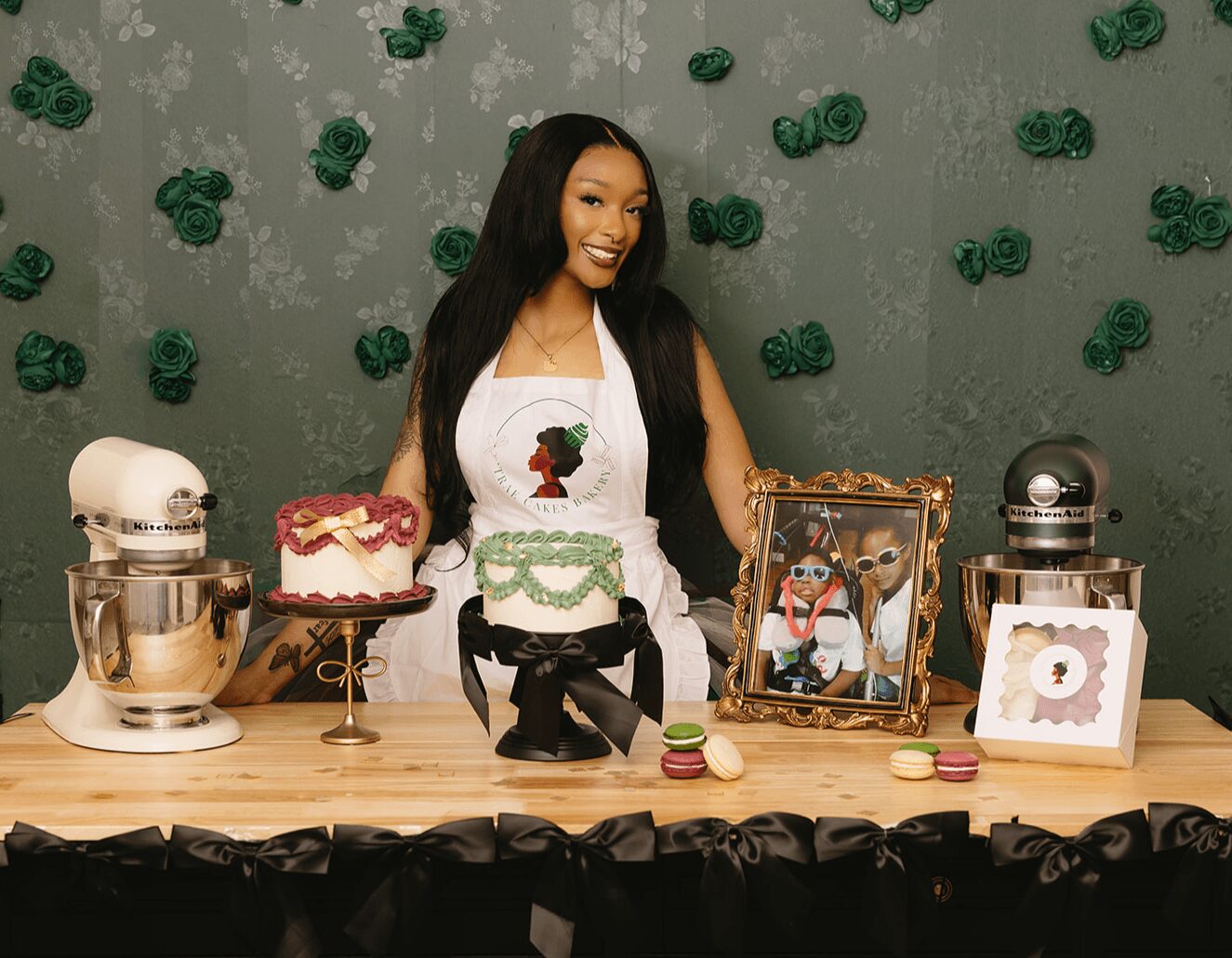 Woman smiling at a decorated table with cakes, framed photo, and baking items, against a green wall with decorations.