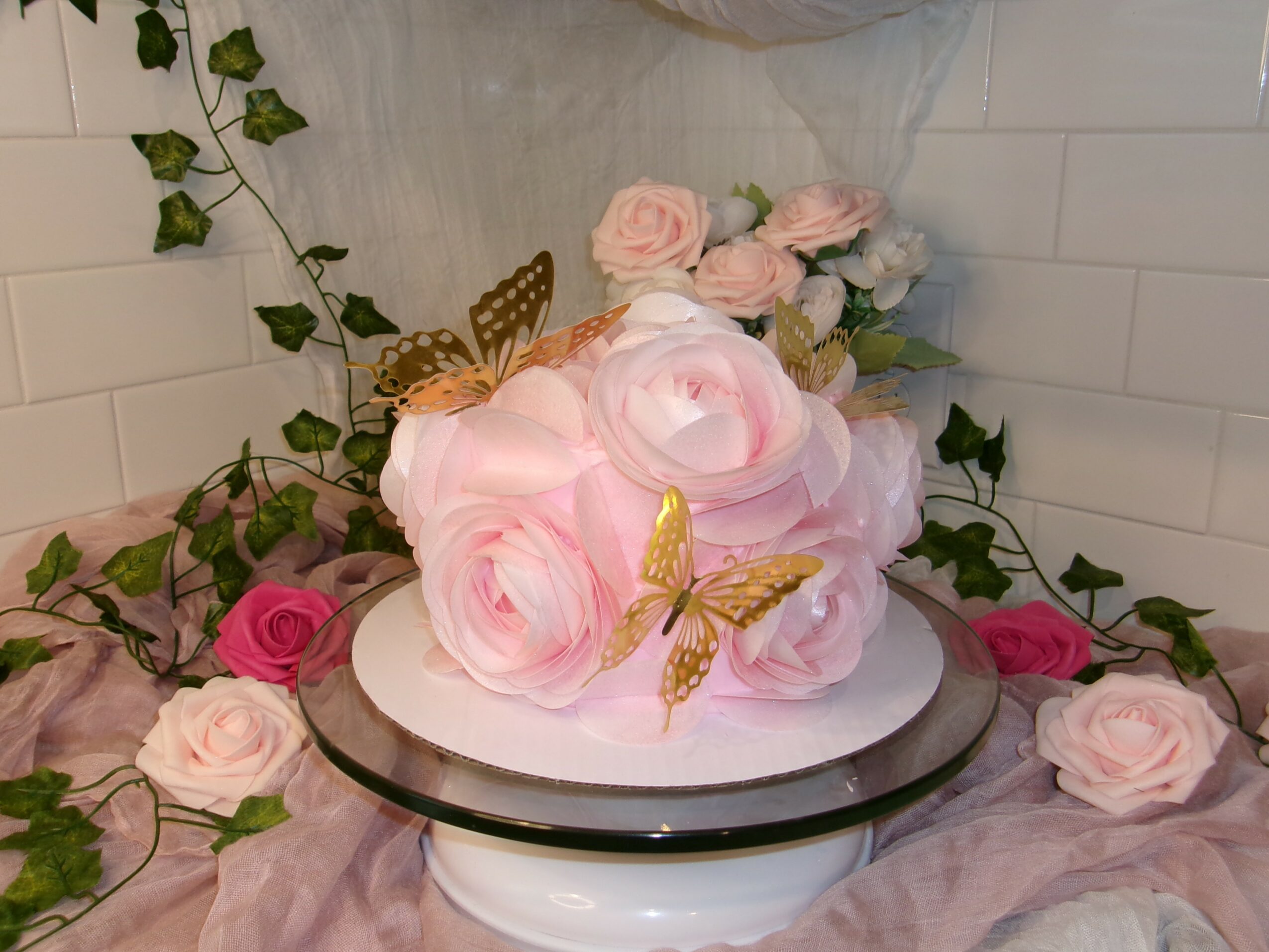 Pink and white roses with gold butterfly decorations on a cake, surrounded by roses and greenery, on a reflective surface.