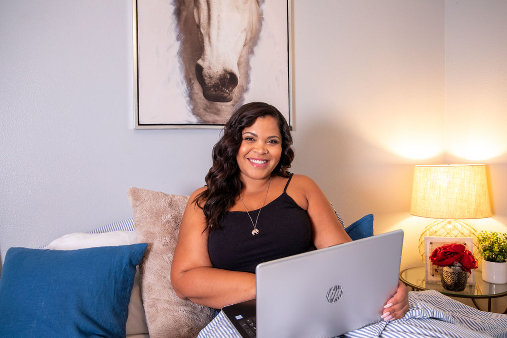 Woman with dark curly hair smiling while sitting at a desk with a laptop, in a cozy room with a horse painting, lamp, and flowers.