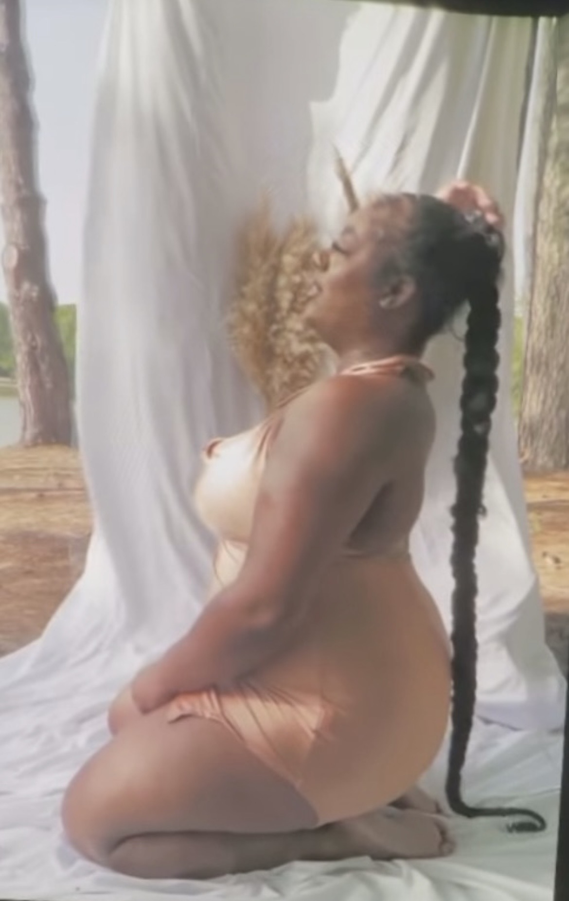 Kneeling woman with long braided hair outdoors, facing a white curtain, with trees in the background.