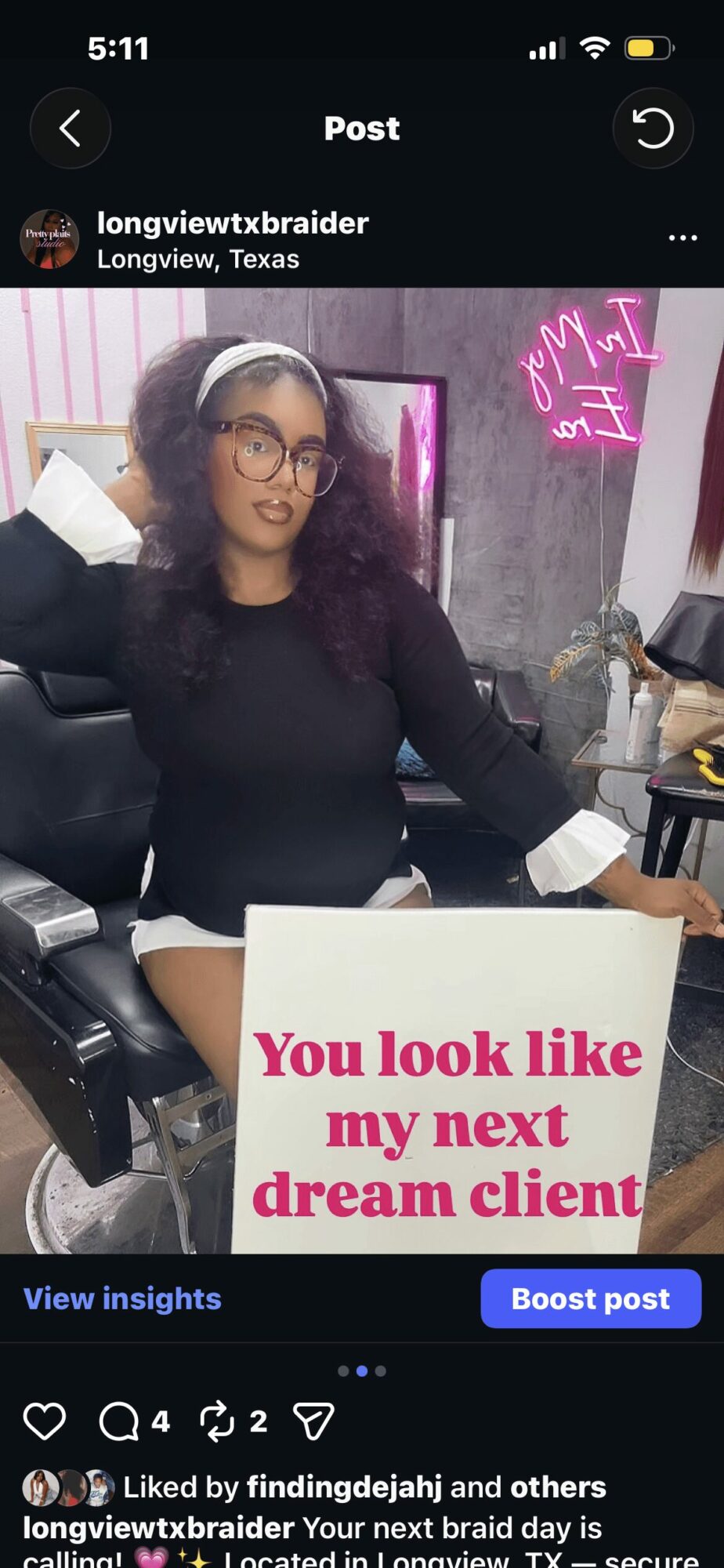 Woman with glasses and curly hair sitting in a chair, wearing a black top and white pants, in a salon or studio setting.