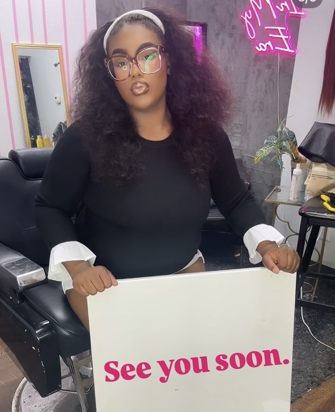 Woman with curly hair and glasses holding a sign that says 'See you soon' in a salon.