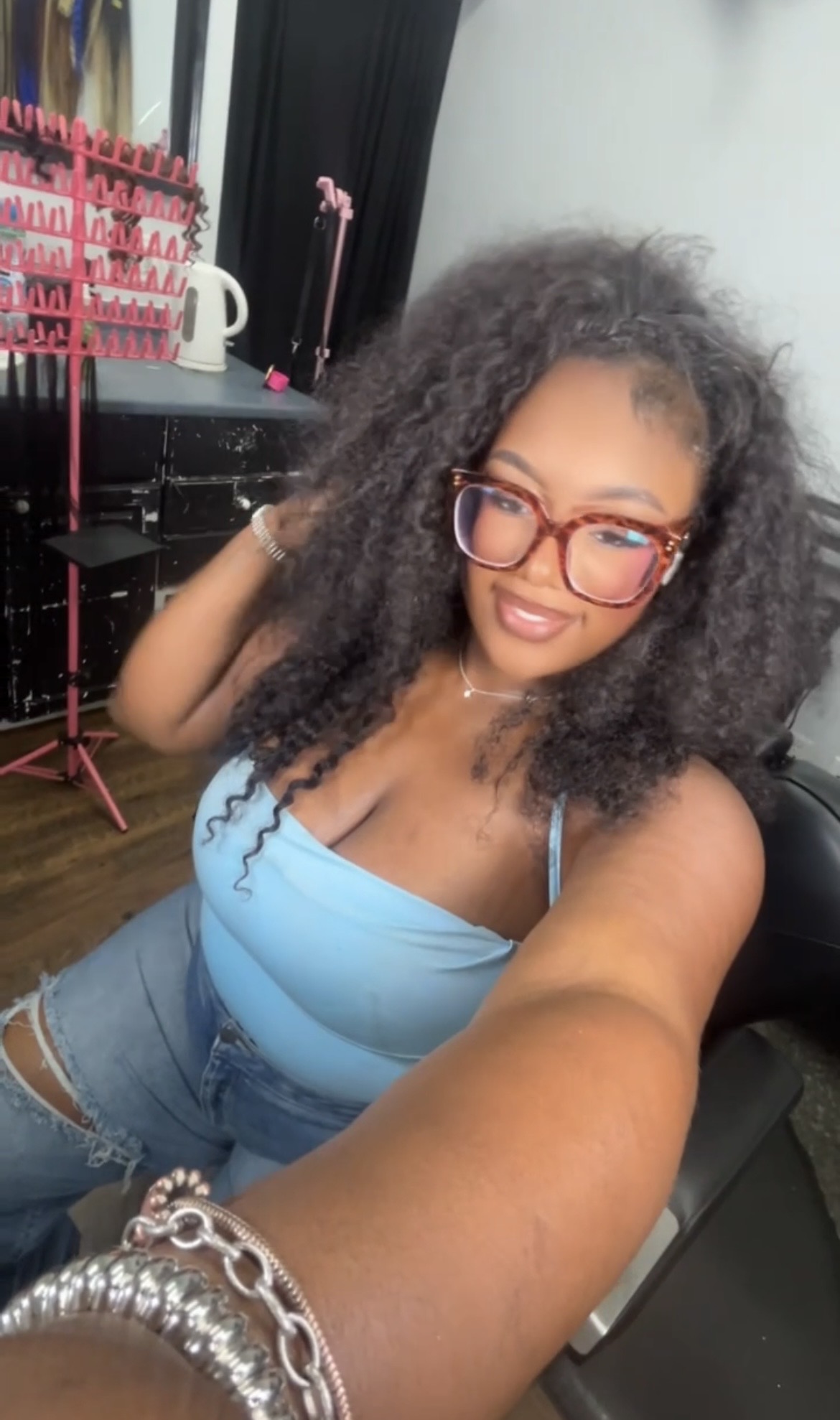 Young woman with curly hair and glasses taking a selfie, wearing a blue top and jeans, in a room with black and red furniture.