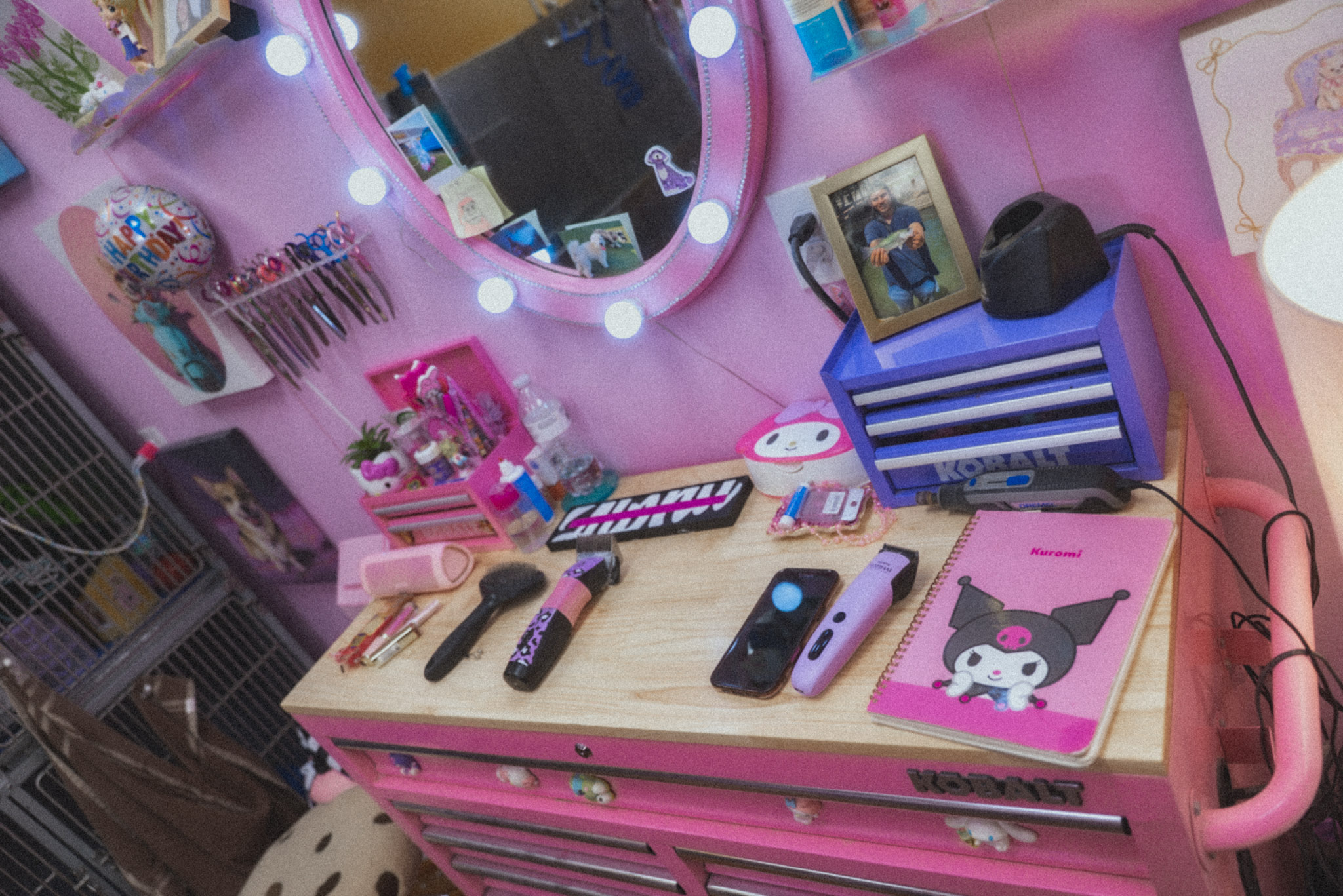 Pink vanity table with mirror, makeup brushes, remote controls, phone, and a pink notebook with a cartoon cat, in a decorated room.