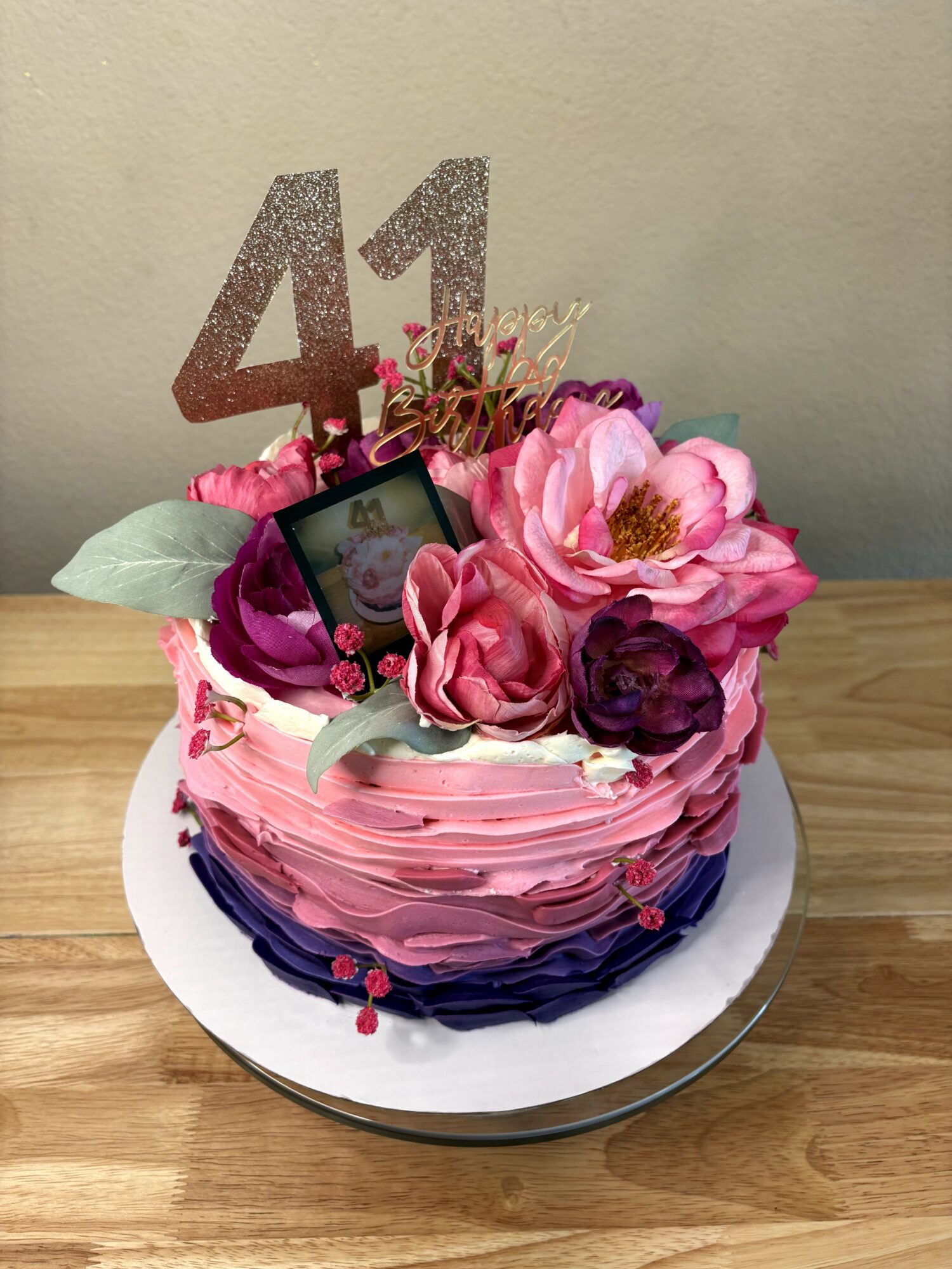 Round birthday cake decorated with pink and purple flowers, topped with a glittery number 41, on a white plate.