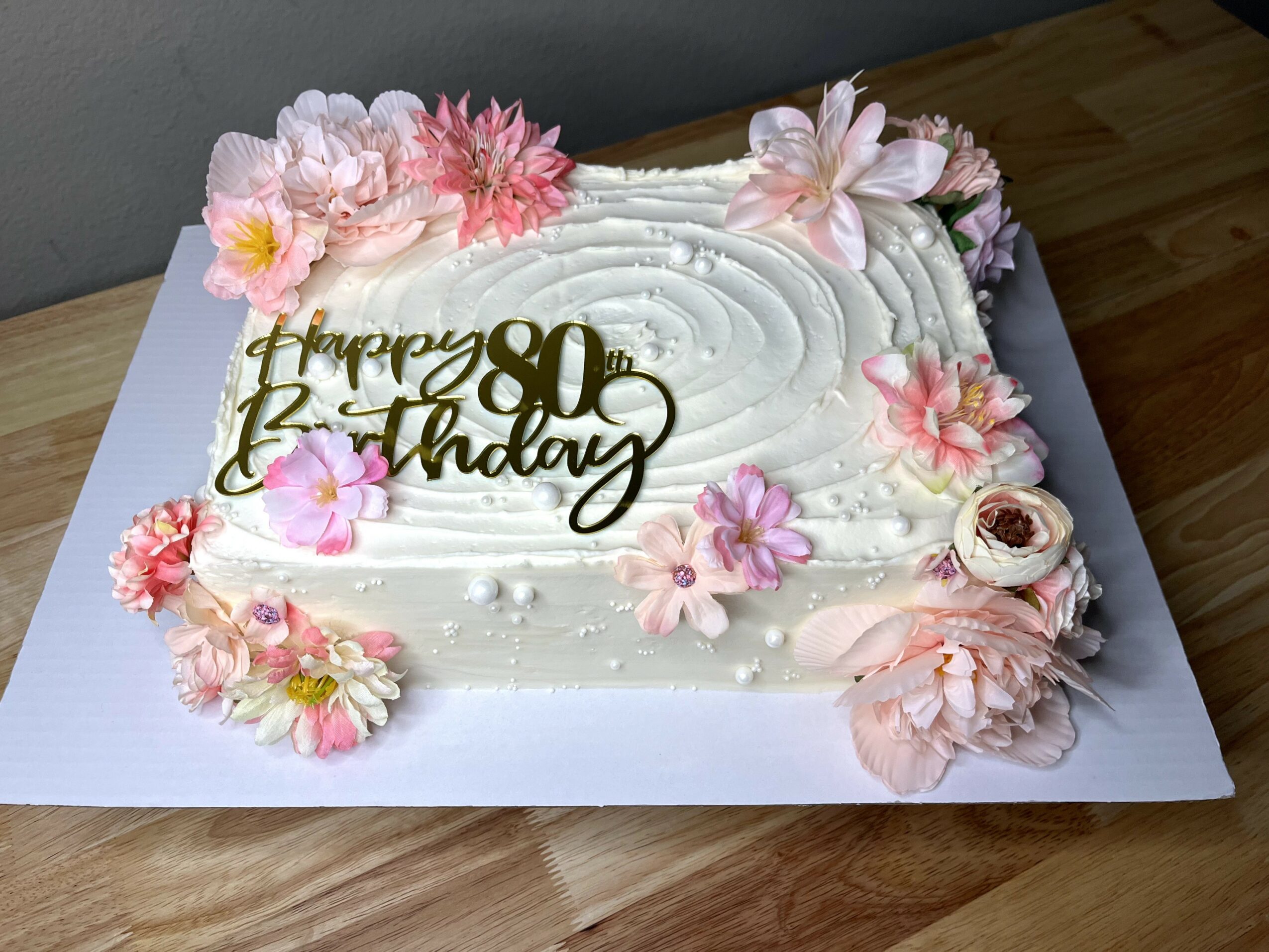 Rectangular birthday cake decorated with pink and white flowers, a gold 'Happy 80th Birthday' topper, on a white base.