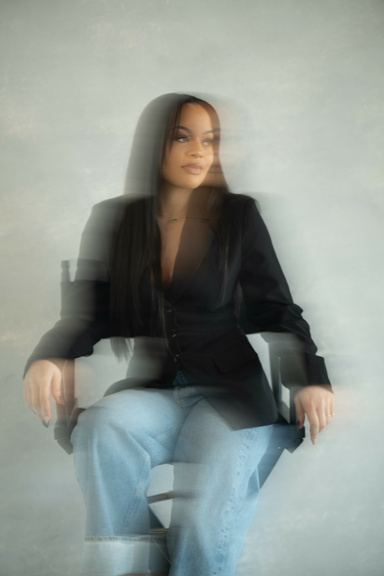 Woman sitting on a chair, wearing a black blazer and light jeans, against a plain background.
