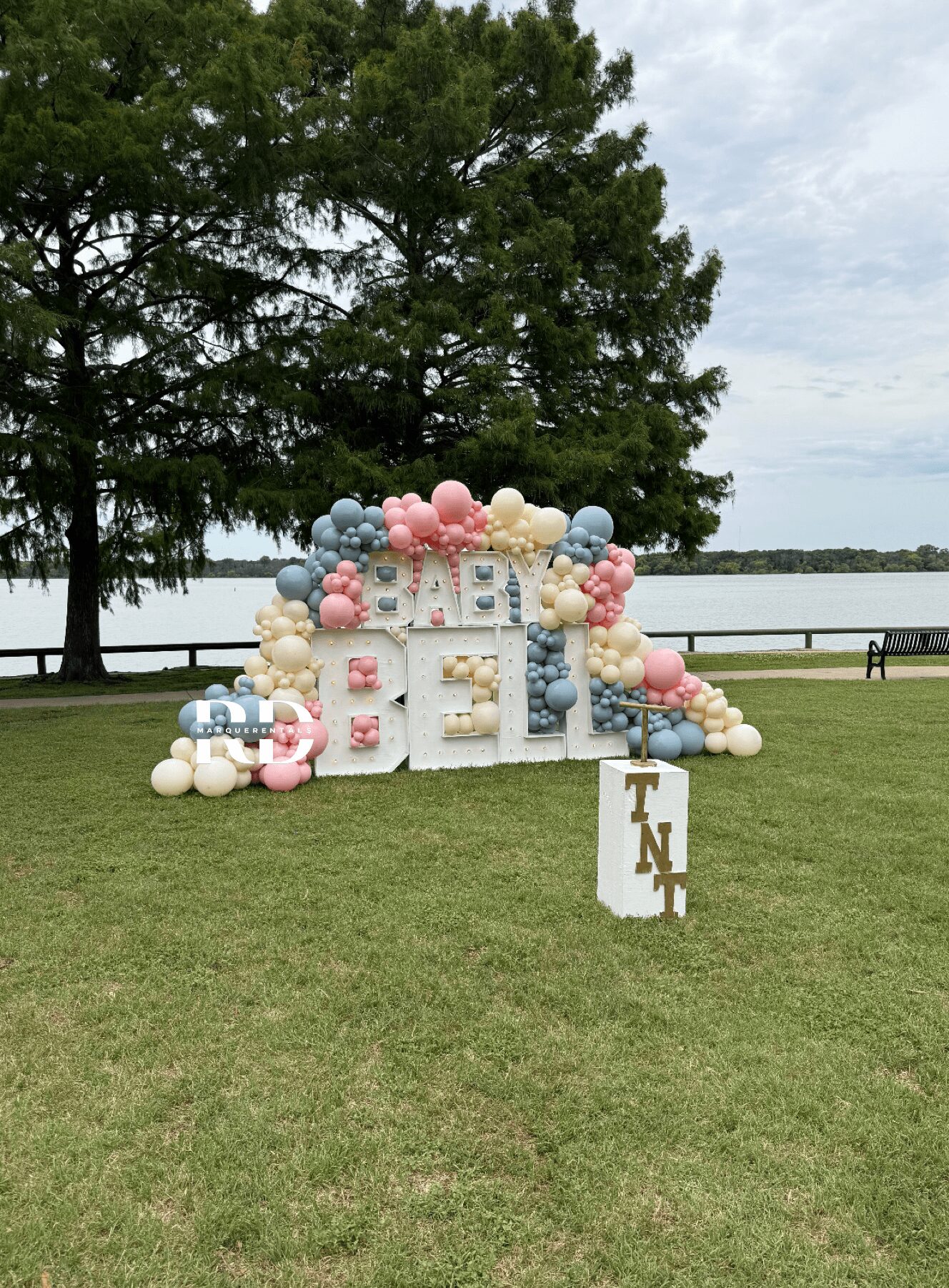 Decorative balloon arch with pastel colors and large blocks spelling 'BABY' on grass near water, with trees and a person in the background.