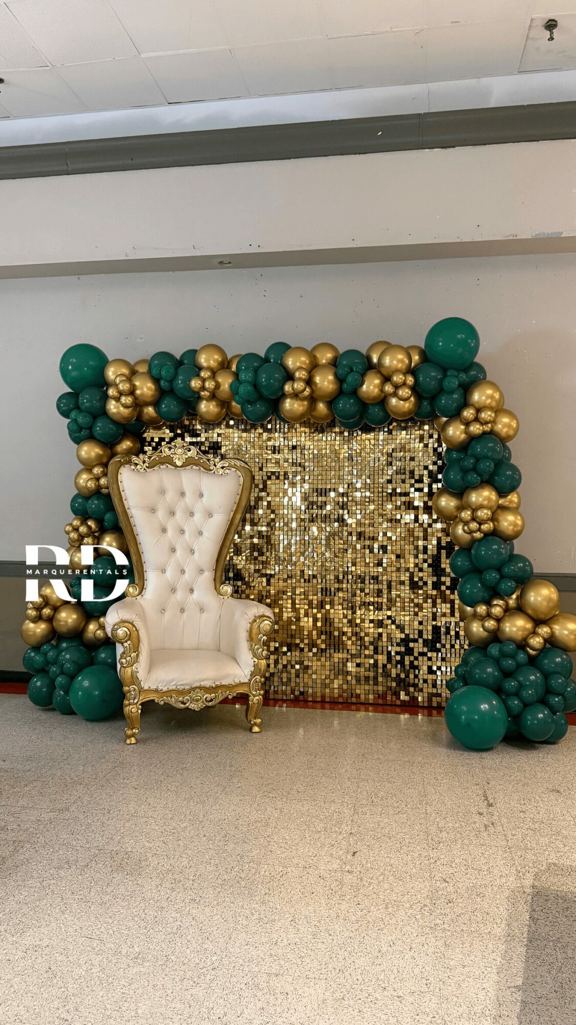Decorative setup with a white ornate chair, gold and green balloons, and a gold sequin backdrop.