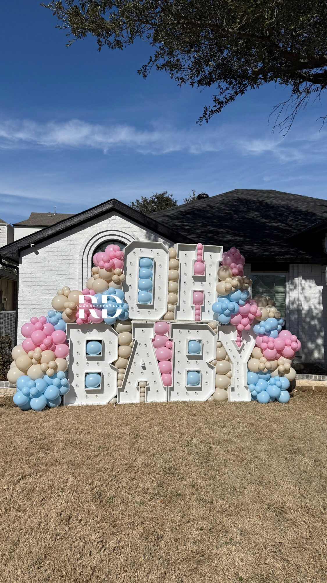 Decorative display with large letters spelling 'BABY' and colorful balloons outdoors, with a house and trees in background.