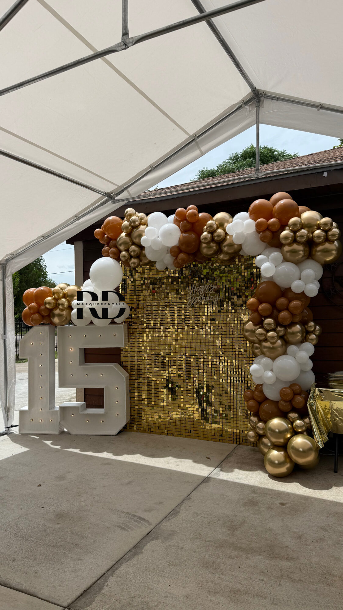 Decorative backdrop with balloons in gold, white, and bronze, and a gold curtain, under a tent at an outdoor event.