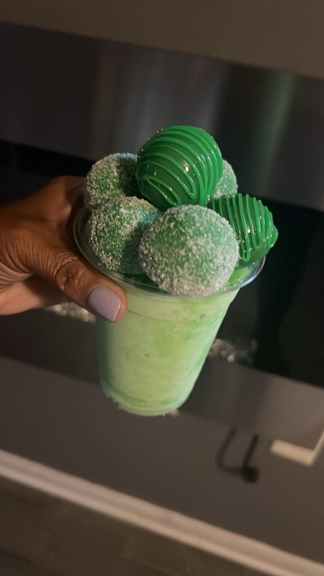 Hand holding a glass of green smoothie topped with green and white spherical candies and green striped chocolates.