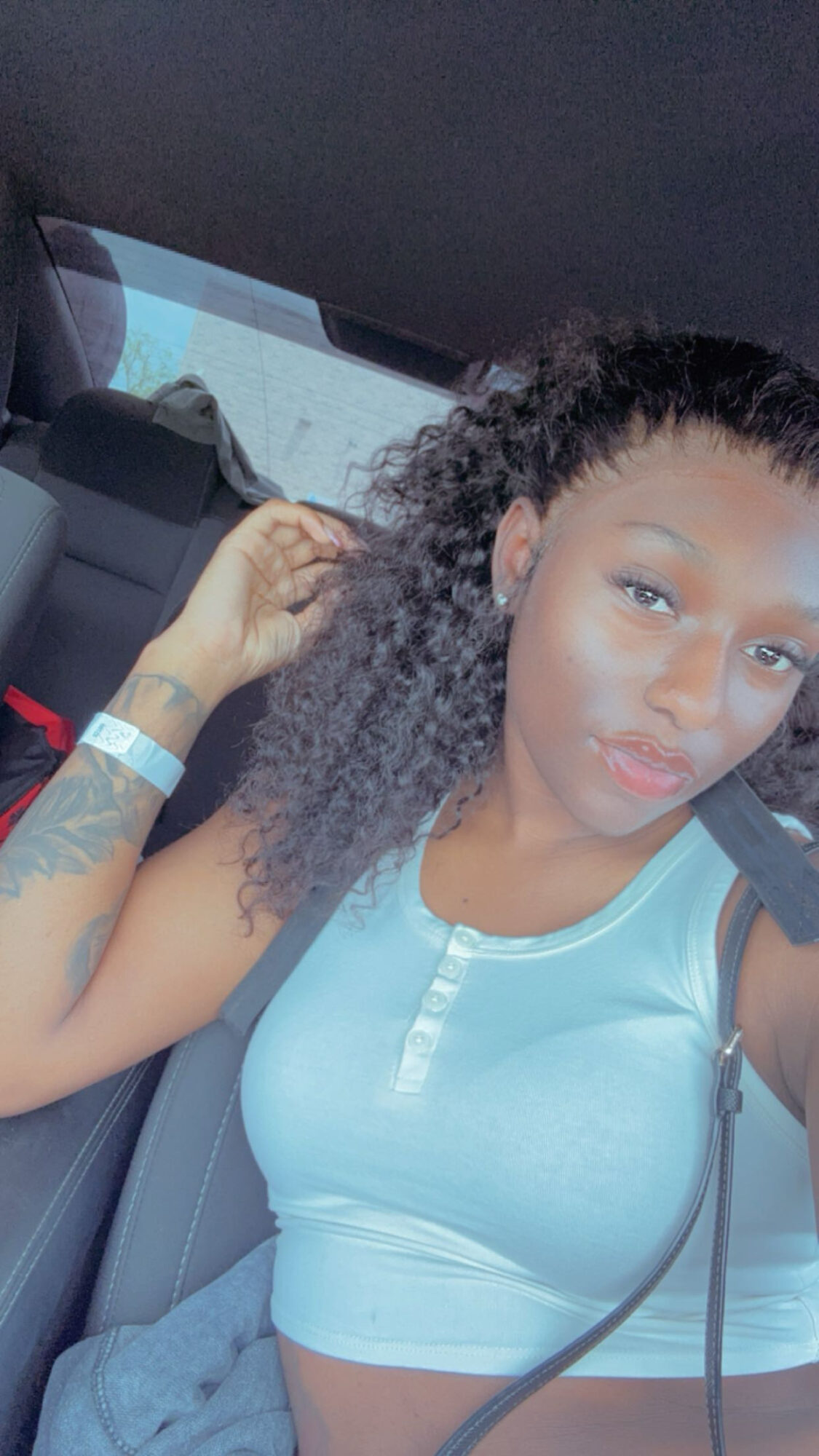 Young woman with curly hair sitting in a car, wearing a light blue sleeveless top, looking at the camera.