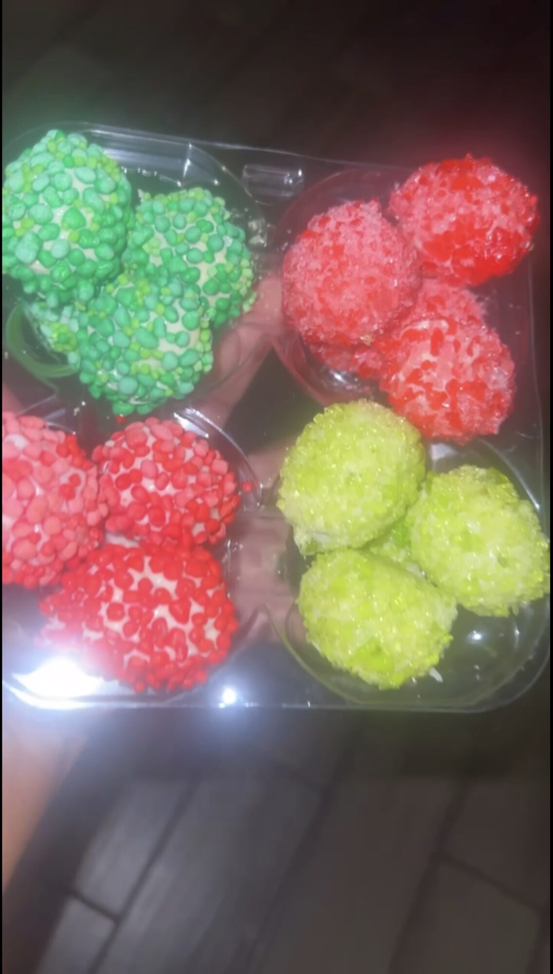 Colorful candy-coated confections in green, red, and yellow, arranged in a plastic container, with a reflective surface below.