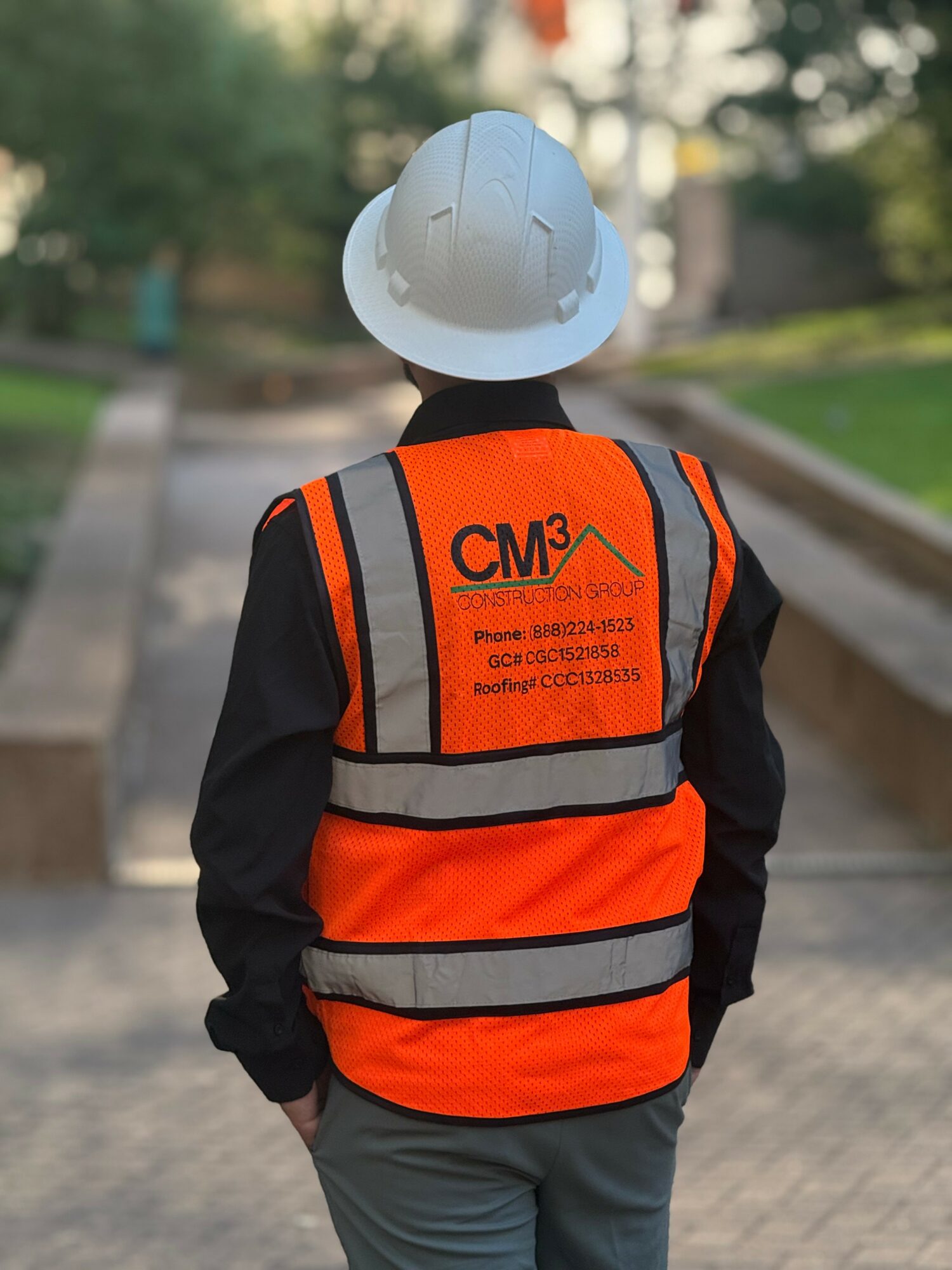 Person wearing a white safety helmet and orange reflective vest walking outdoors on a paved path, with trees in the background.