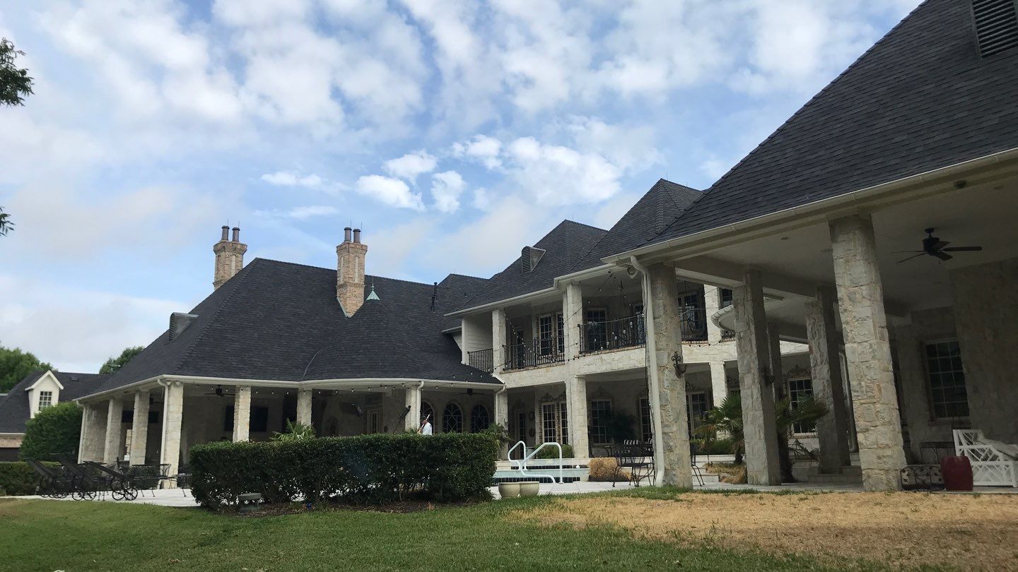 Large house with multiple chimneys, covered porch, and a balcony, set against a partly cloudy sky.