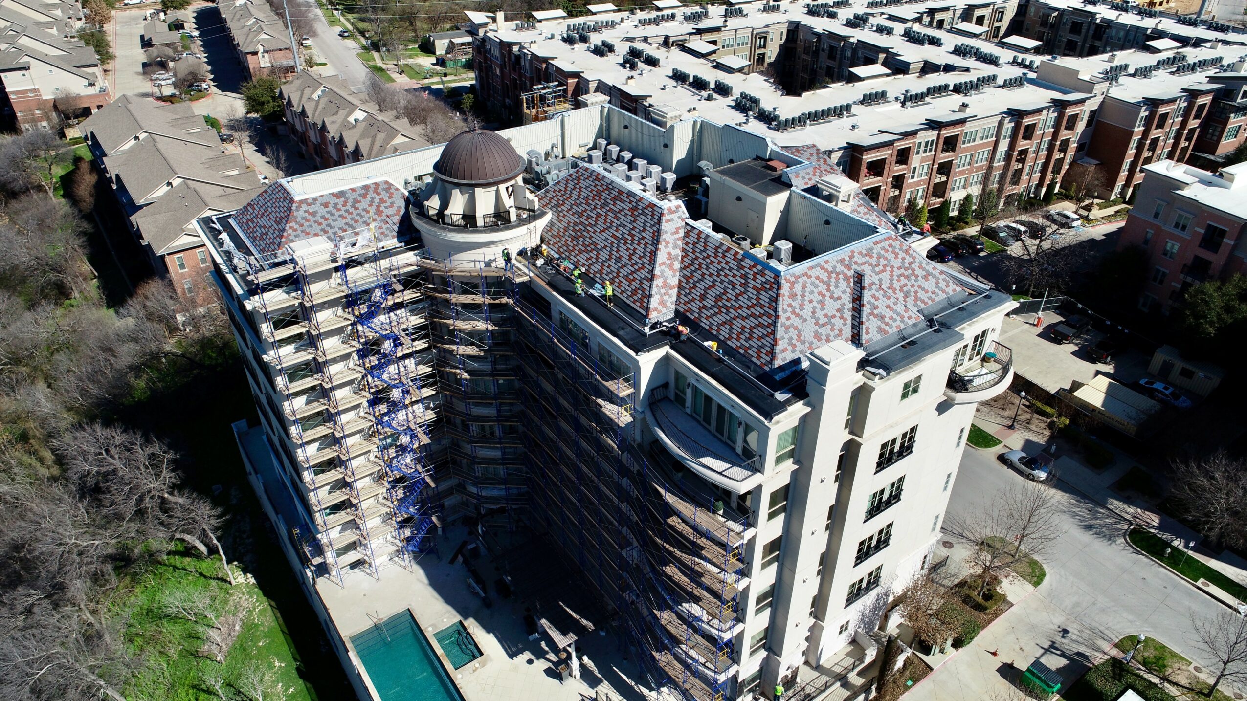 A large multi-story building with a rounded rooftop structure, surrounded by trees and neighboring buildings, viewed from above.