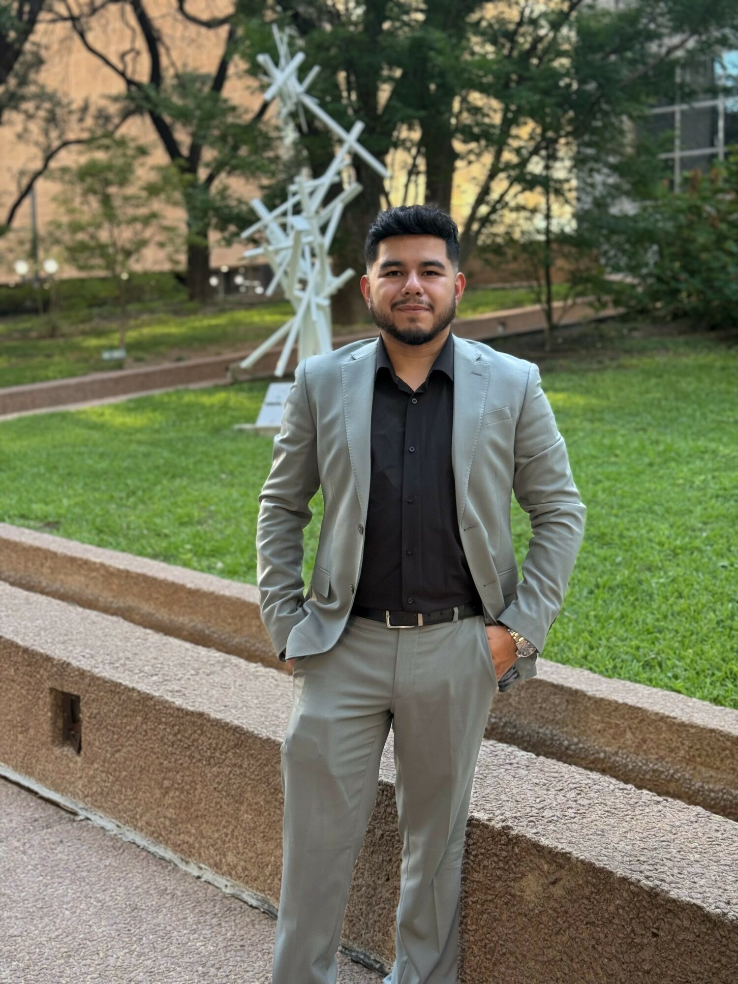 Young man in a light gray suit and black shirt standing outdoors on a grassy area with trees and modern art sculpture behind him.