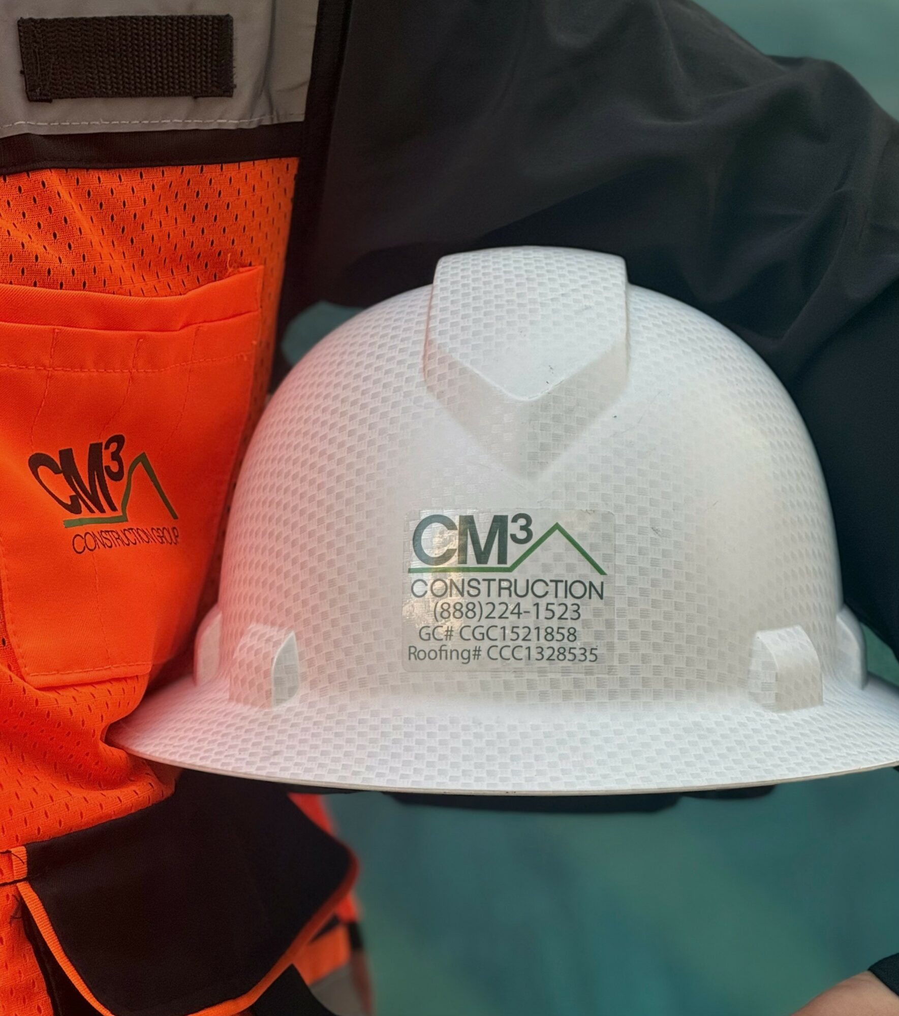 Person holding a white construction helmet with company logo and contact information, wearing a black and orange safety vest.