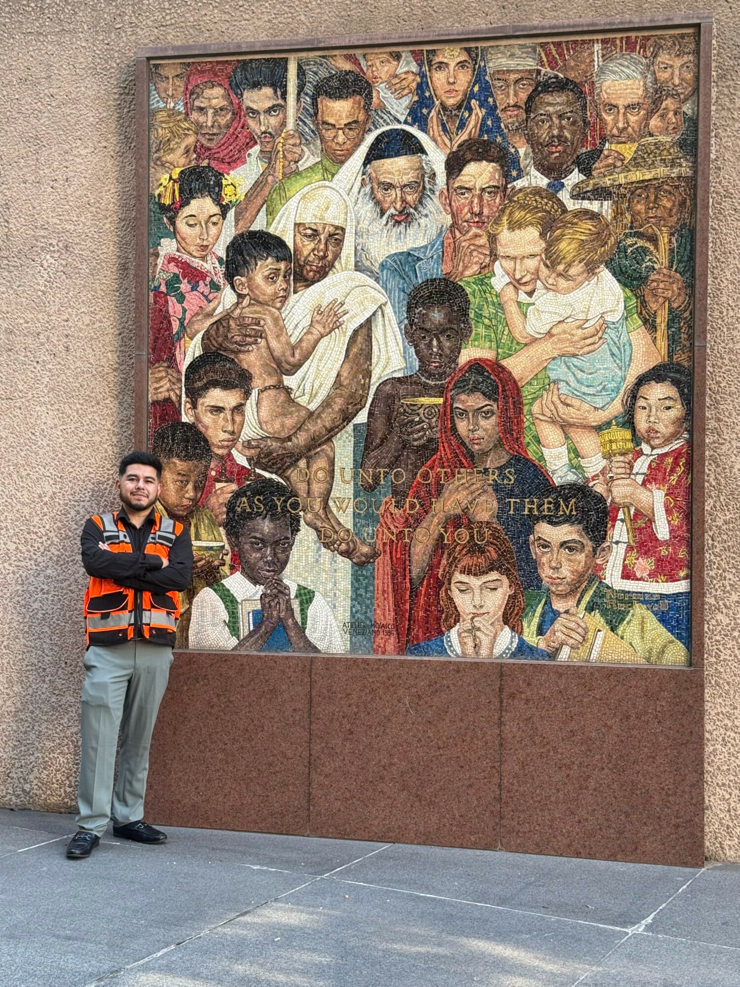 Person standing next to a large colorful mural depicting diverse people, including children and adults, in a crowded scene.