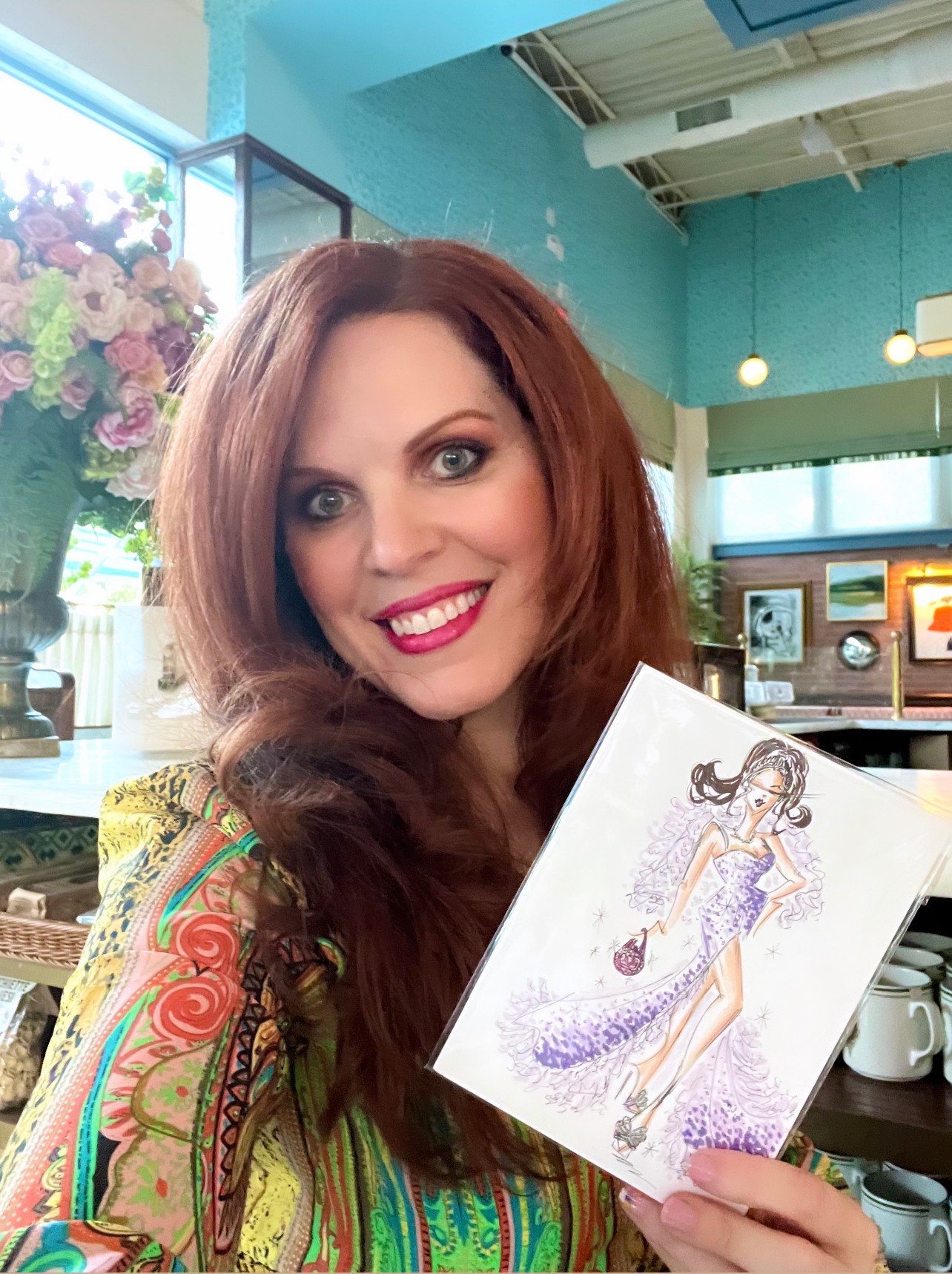 Woman with long red hair smiling, holding a drawing of a woman in a purple dress, inside a colorful cafe.