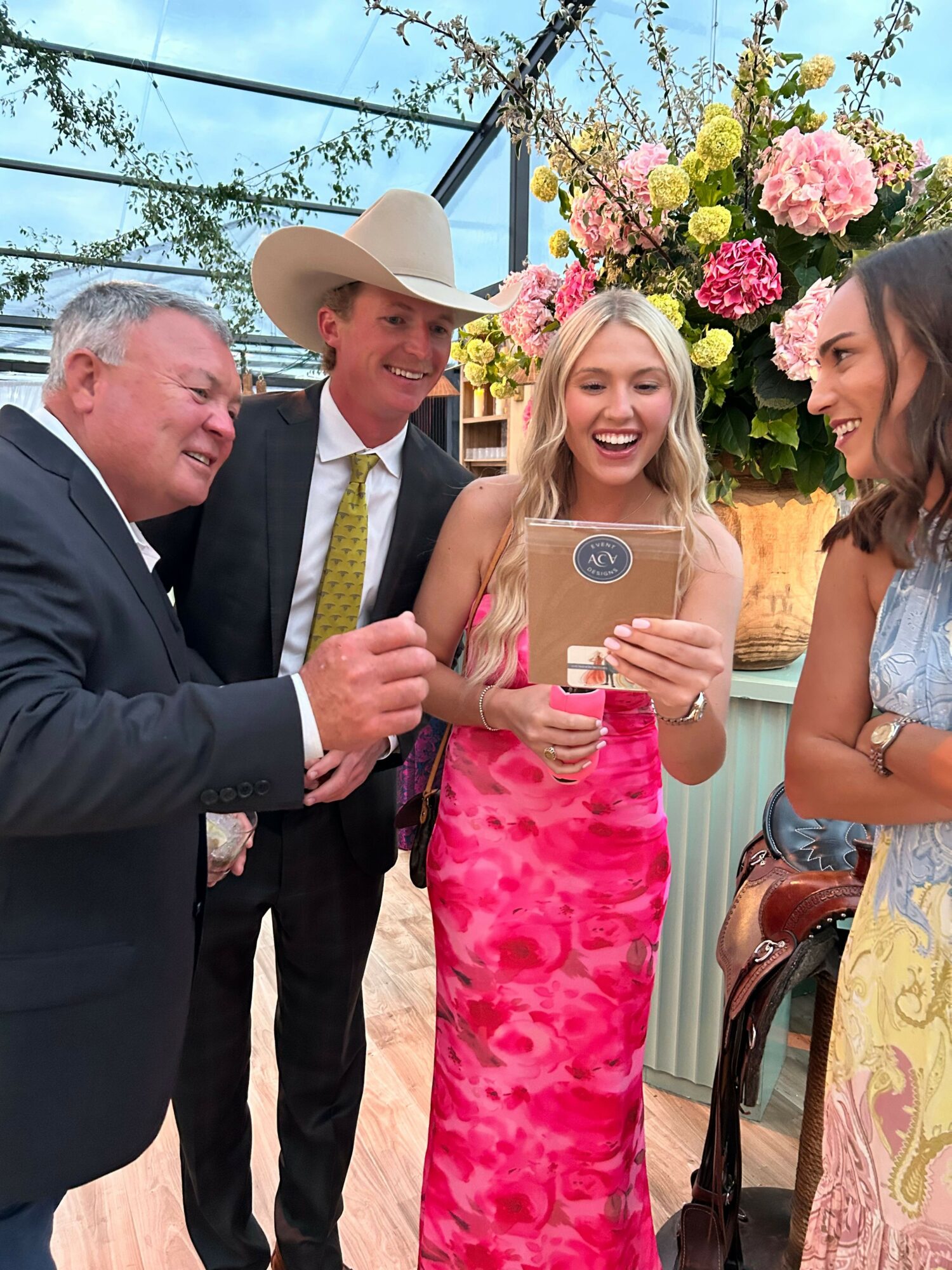 Four people, two women and two men, are outdoors with pink flowers and trees in the background. One woman is taking a selfie.