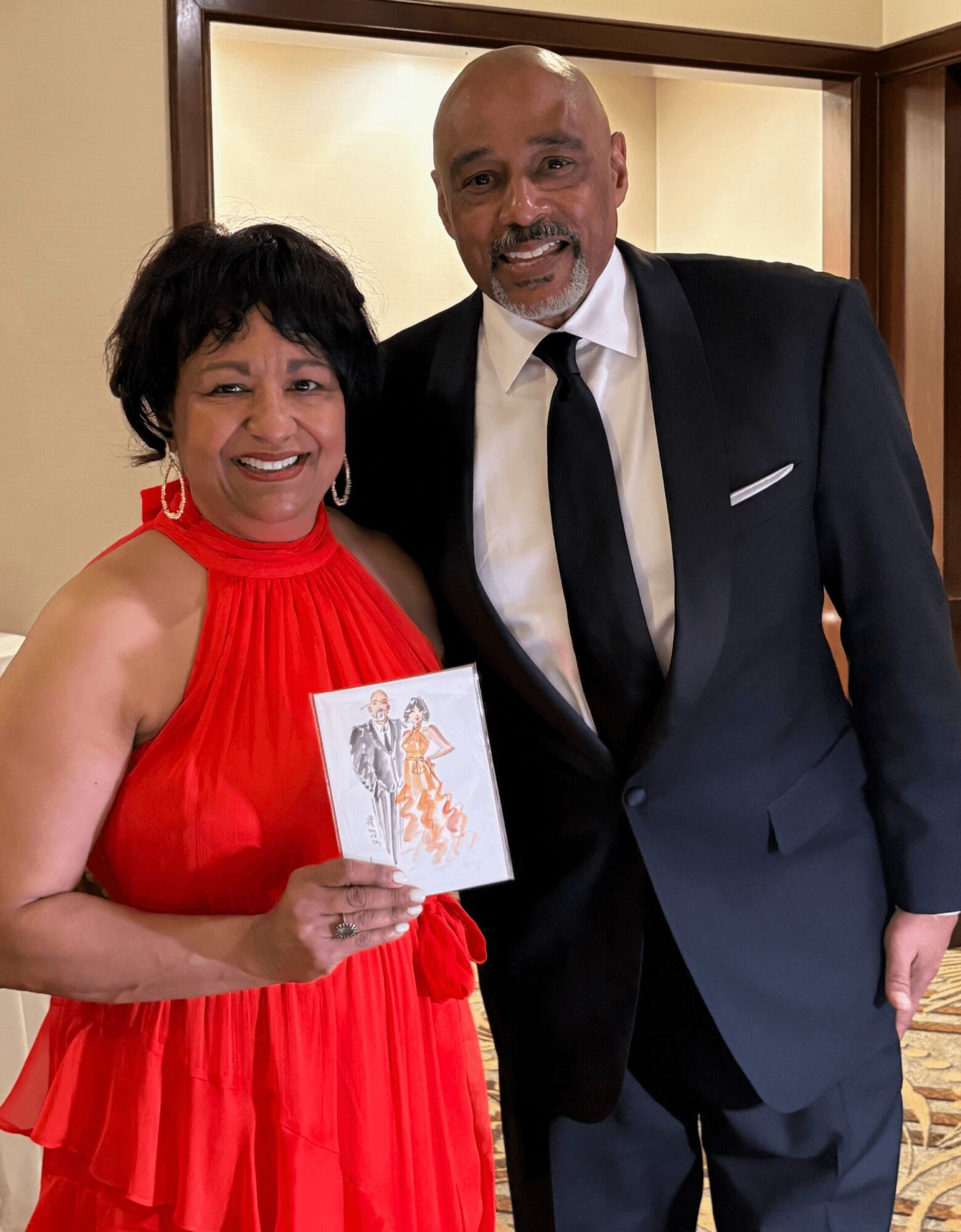 A woman in a red dress holding a card, standing next to a man in a black suit and tie, smiling.