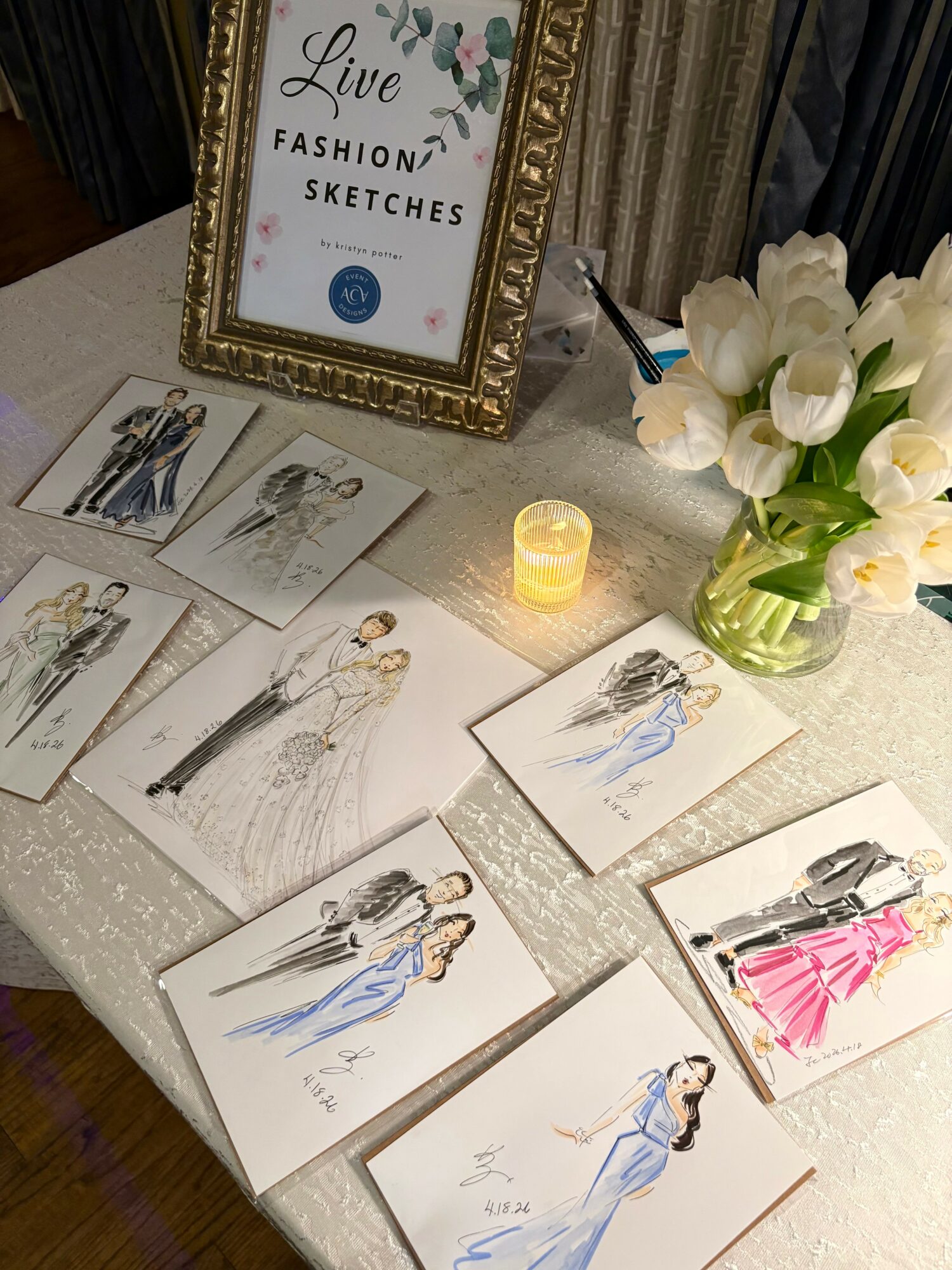 Table displays fashion sketches, a framed sign, a candle, and a vase with white flowers.