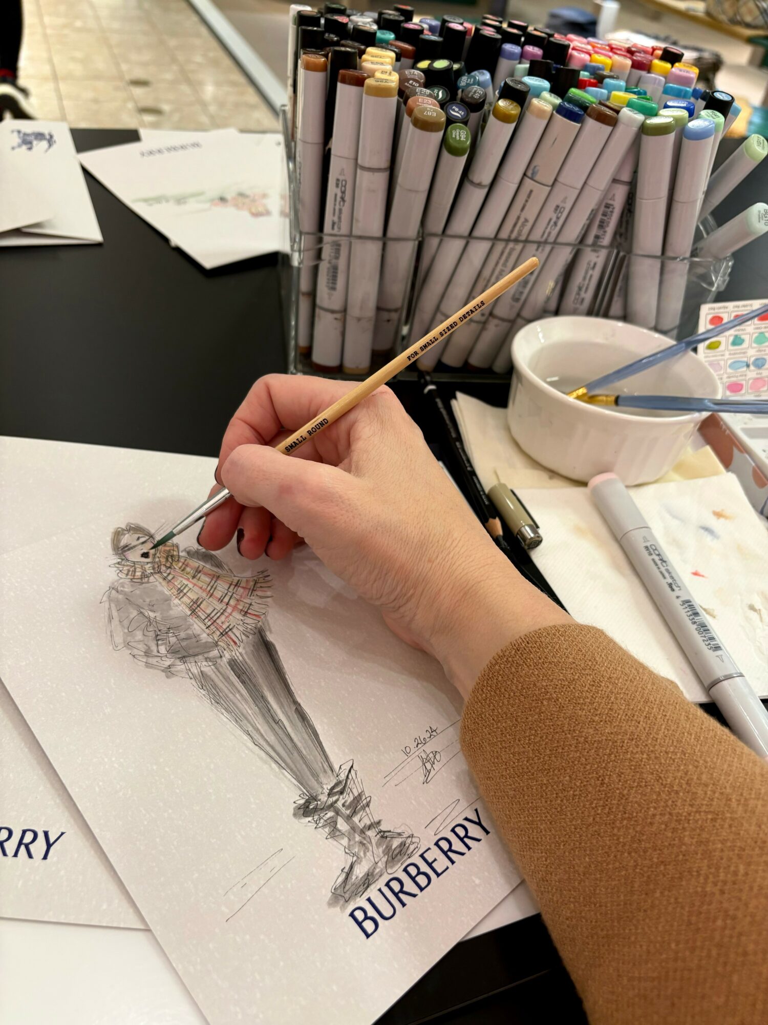 Person coloring a detailed fashion illustration with markers, surrounded by art supplies and a Burberry paper, on a desk.
