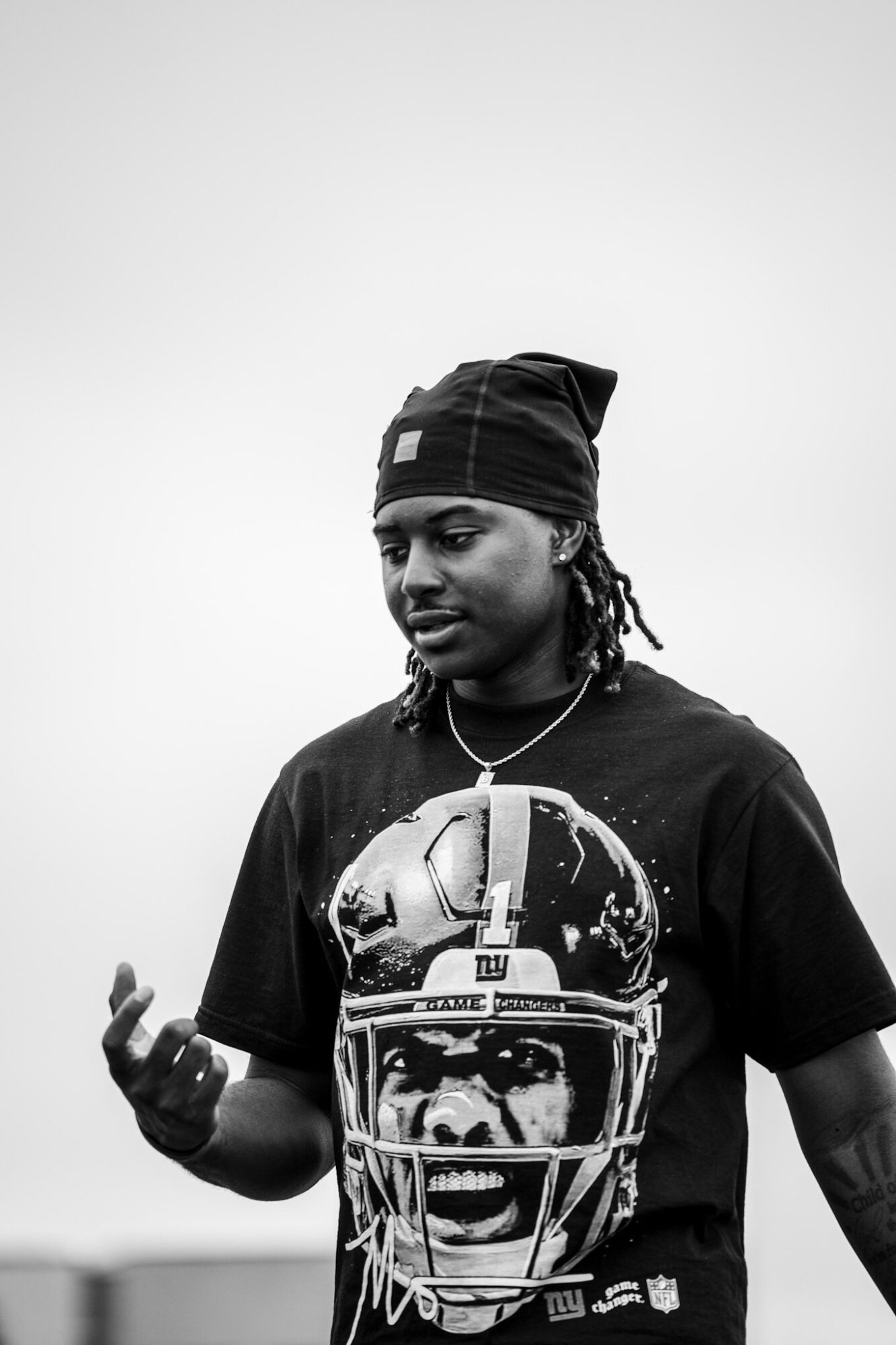 Young man wearing a beanie and a graphic t-shirt, looking down with a neutral expression, against a plain background.