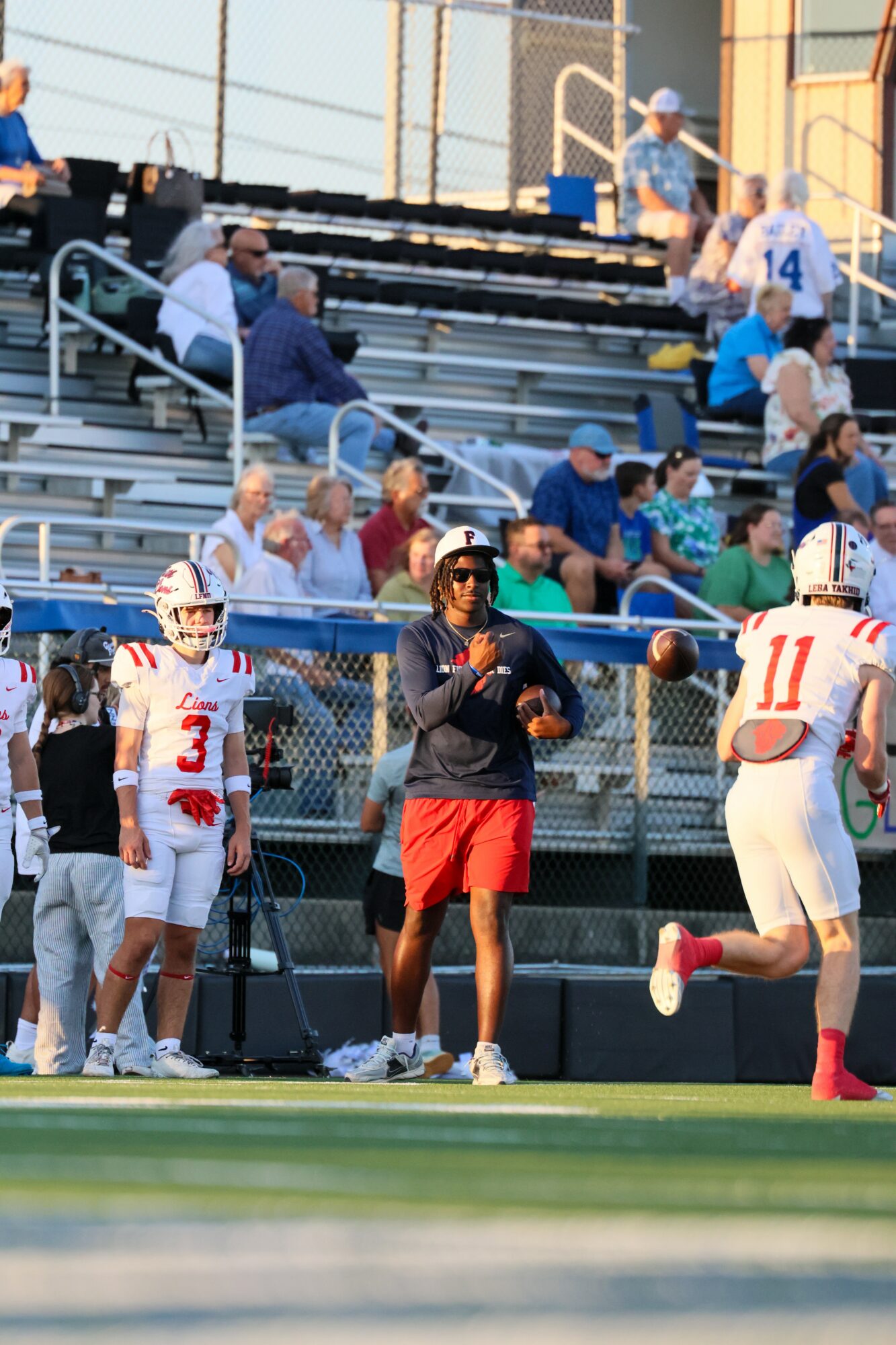 Football players and a coach on the sideline during a game, with spectators in the stands behind a chain-link fence.