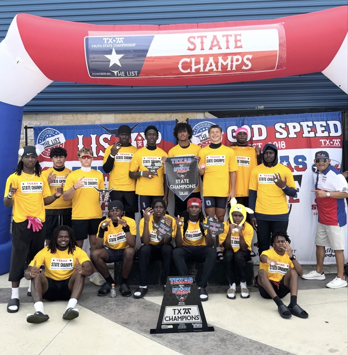 Group of young athletes in yellow shirts holding trophies, celebrating state championship victory outdoors, with banners and an arch in the background.