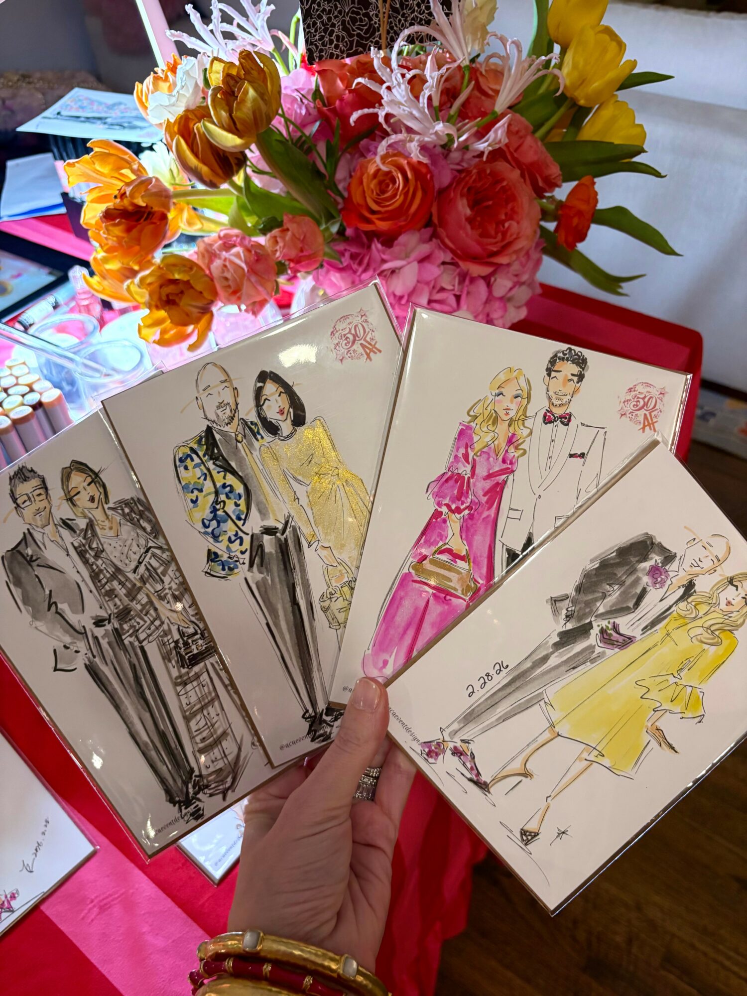 Hand holding four fashion sketches with a colorful flower bouquet in the background.
