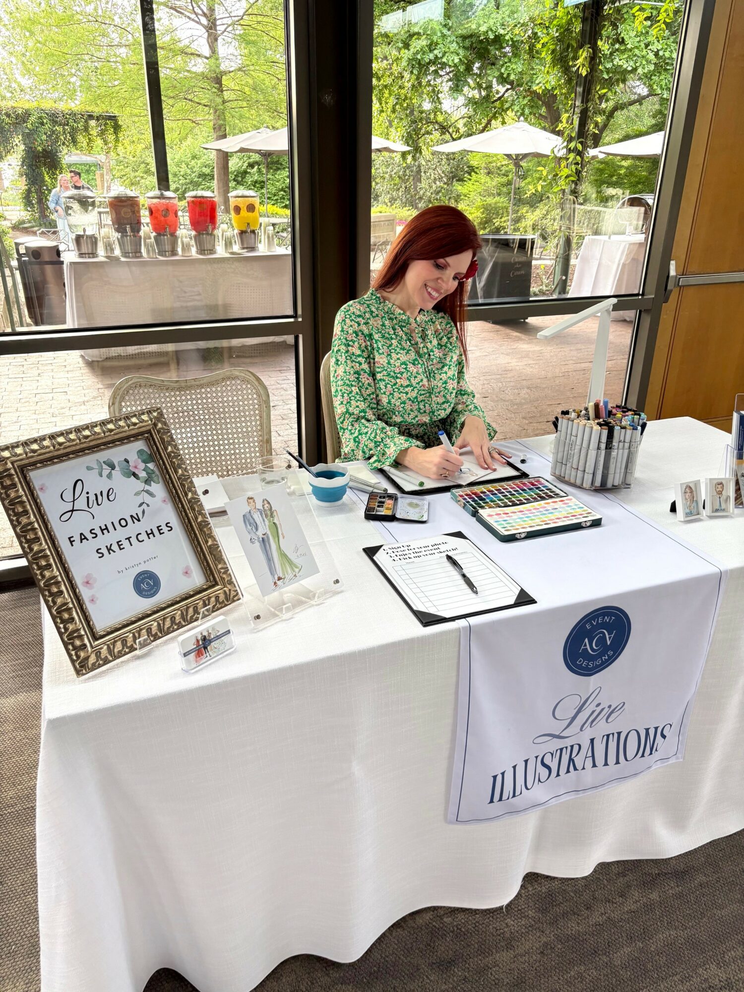 Woman sitting at a table with art supplies, window view of trees and umbrellas outside, sign reading 'Live Illustrations'.