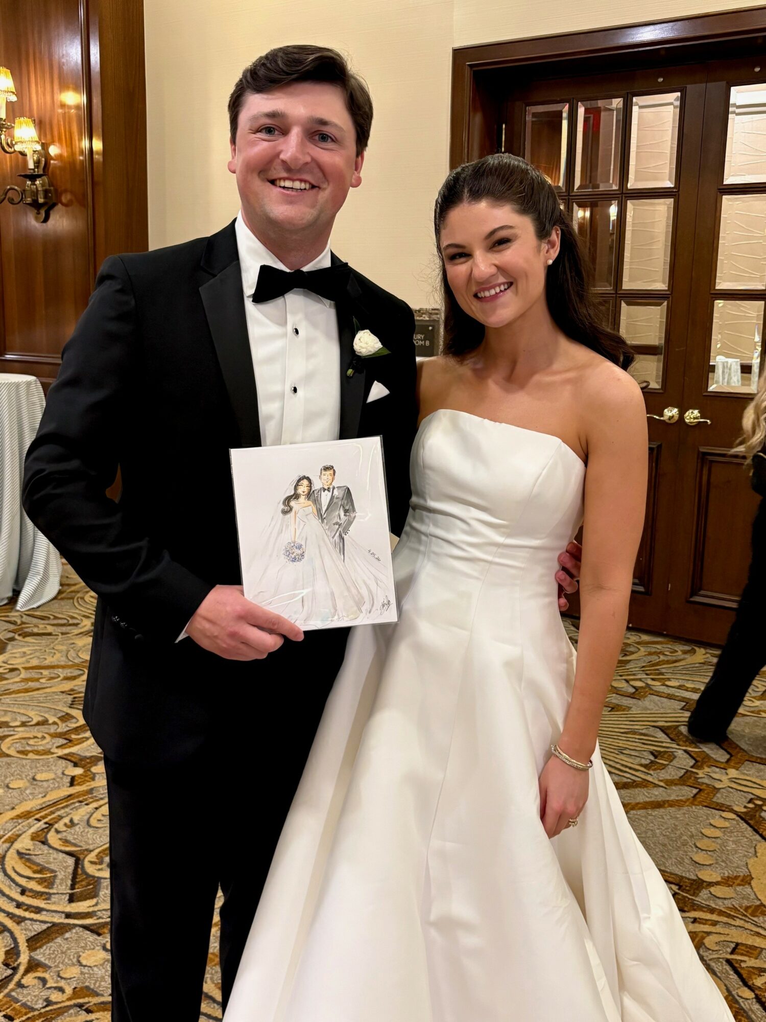 A smiling man in a tuxedo and a woman in a white wedding dress stand together indoors, holding a drawing, with warm lighting and wooden decor.