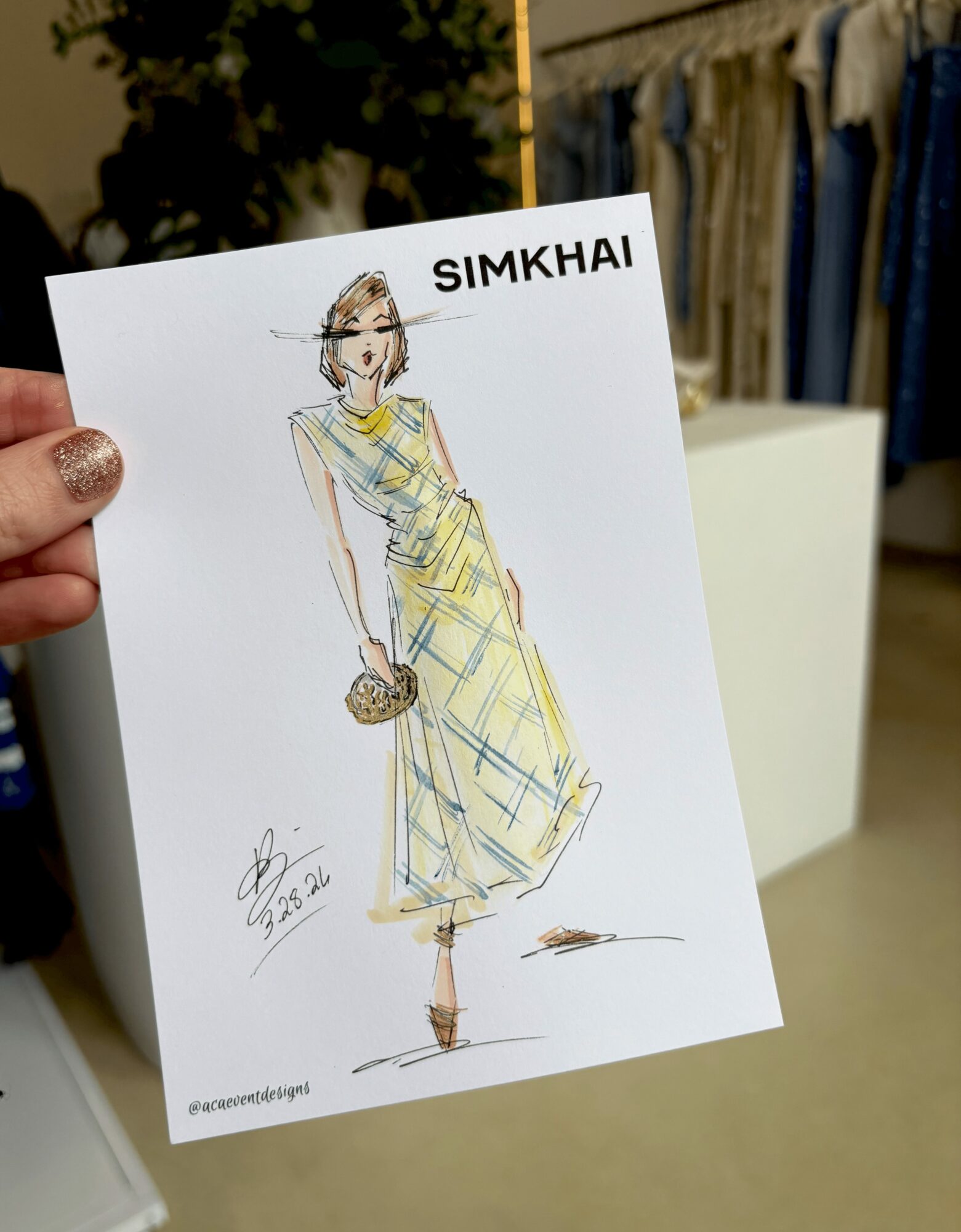 Hand holding a fashion illustration of a woman in a yellow dress with a clutch, in a store setting.