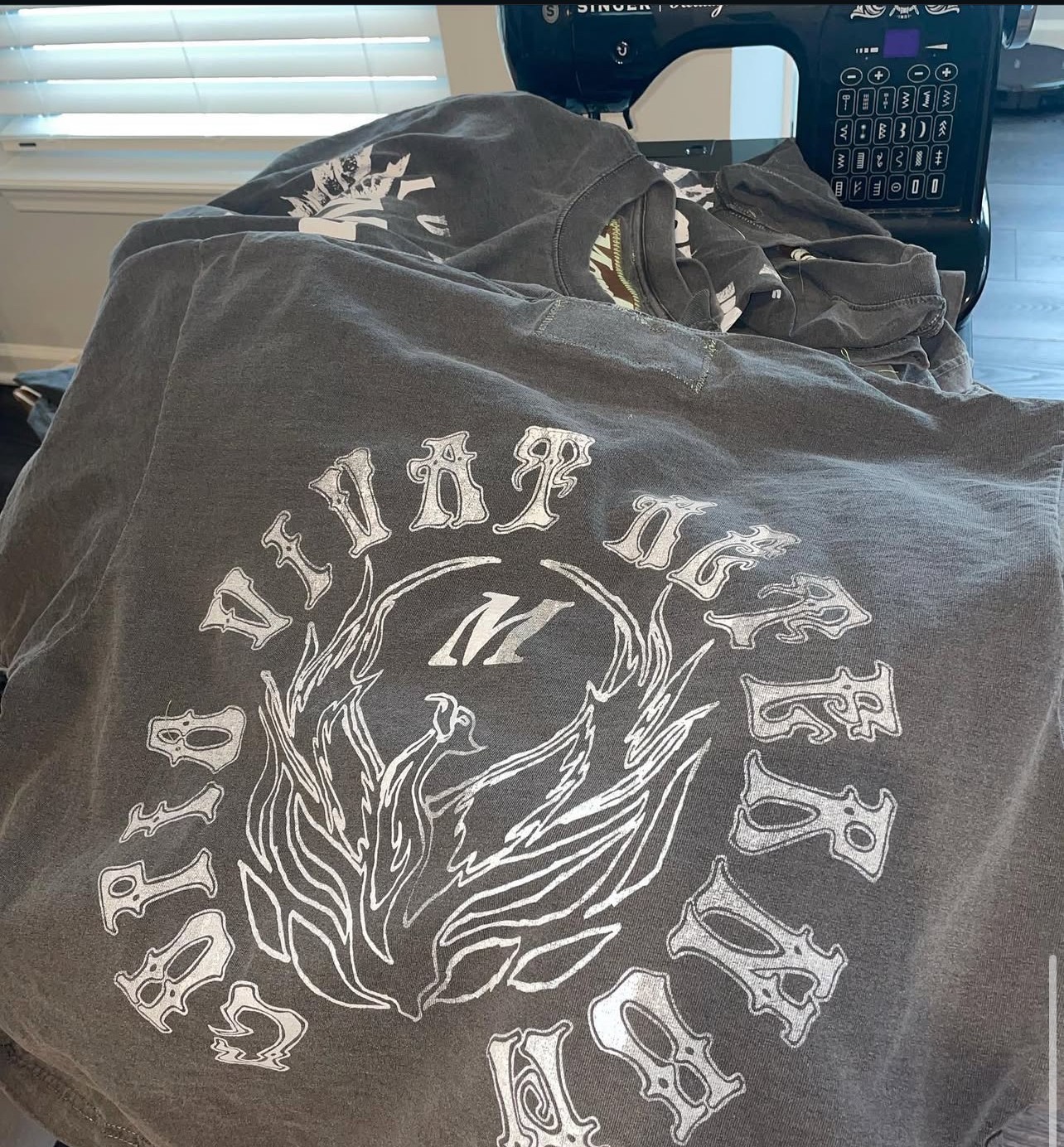 Clothing with a phoenix design and text, placed on a desk near a phone and window blinds.